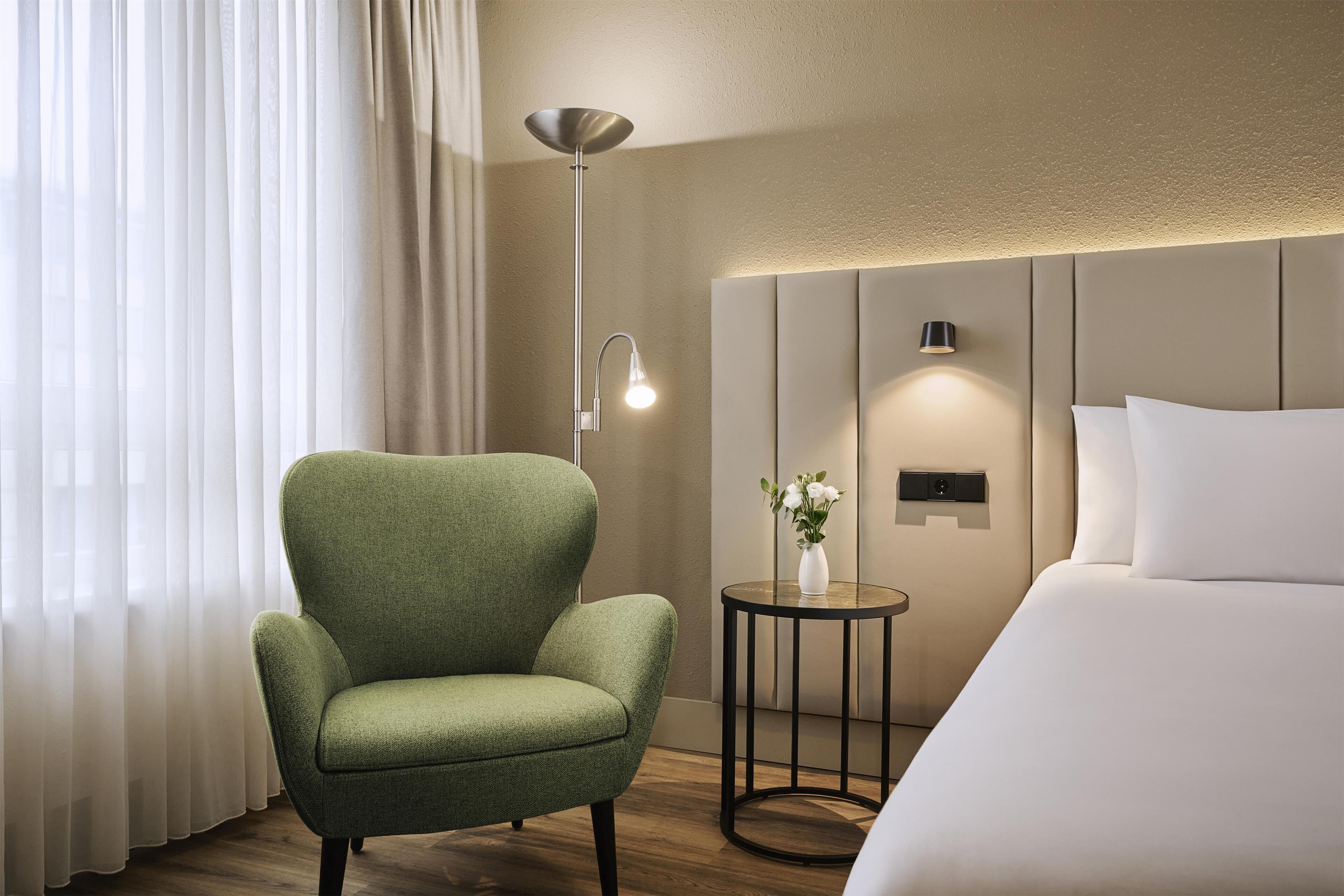 Una habitación de hotel con un sillón verde, una mesita de noche con un jarrón de flores, una lámpara moderna y una cama grande.