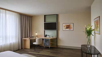 A hotel room with a bed, a desk with a chair, a TV, a small table with a vase, and a window with curtains.