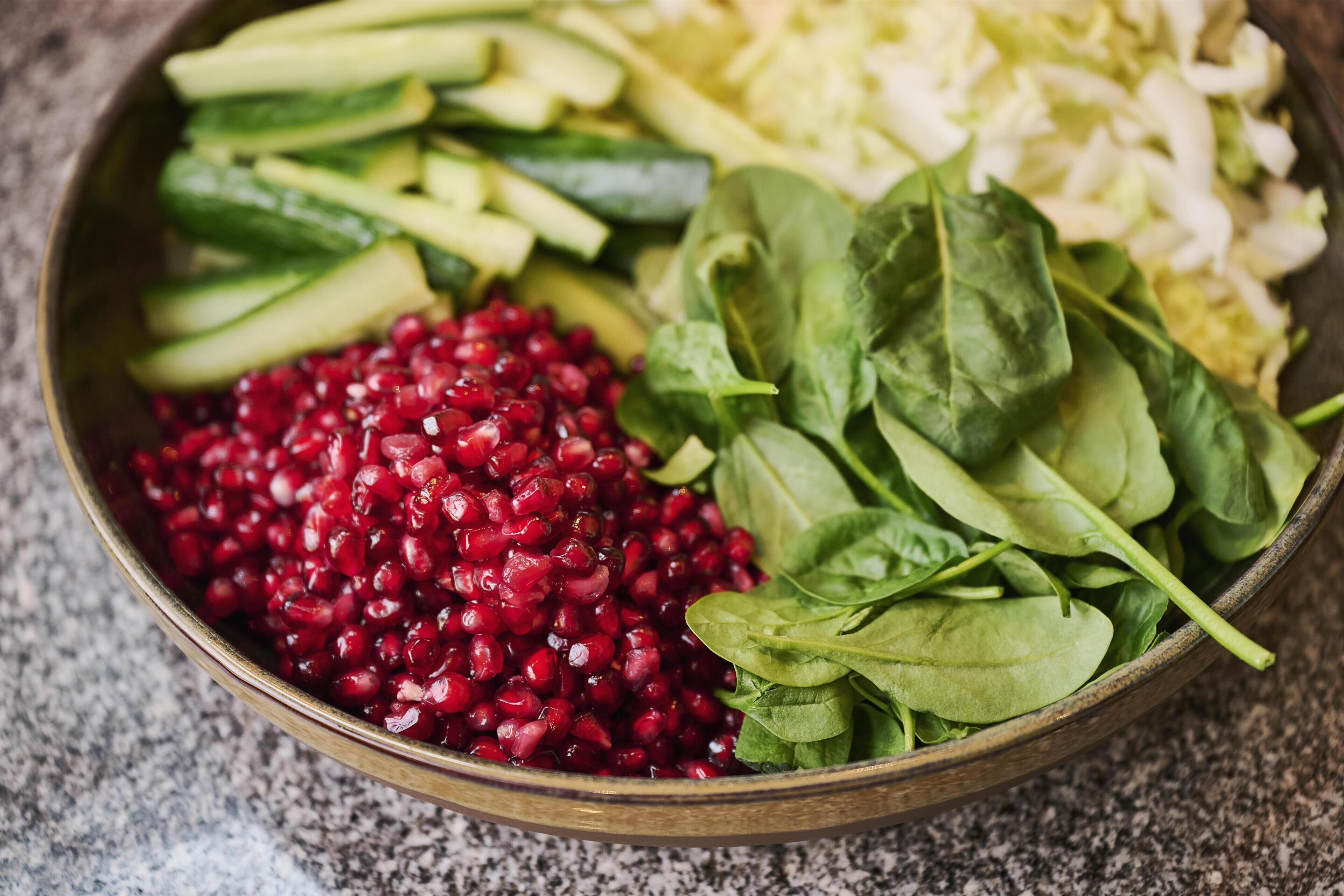 A bowl containing sliced cucumbers, shredded cabbage, spinach leaves, and pomegranate seeds.