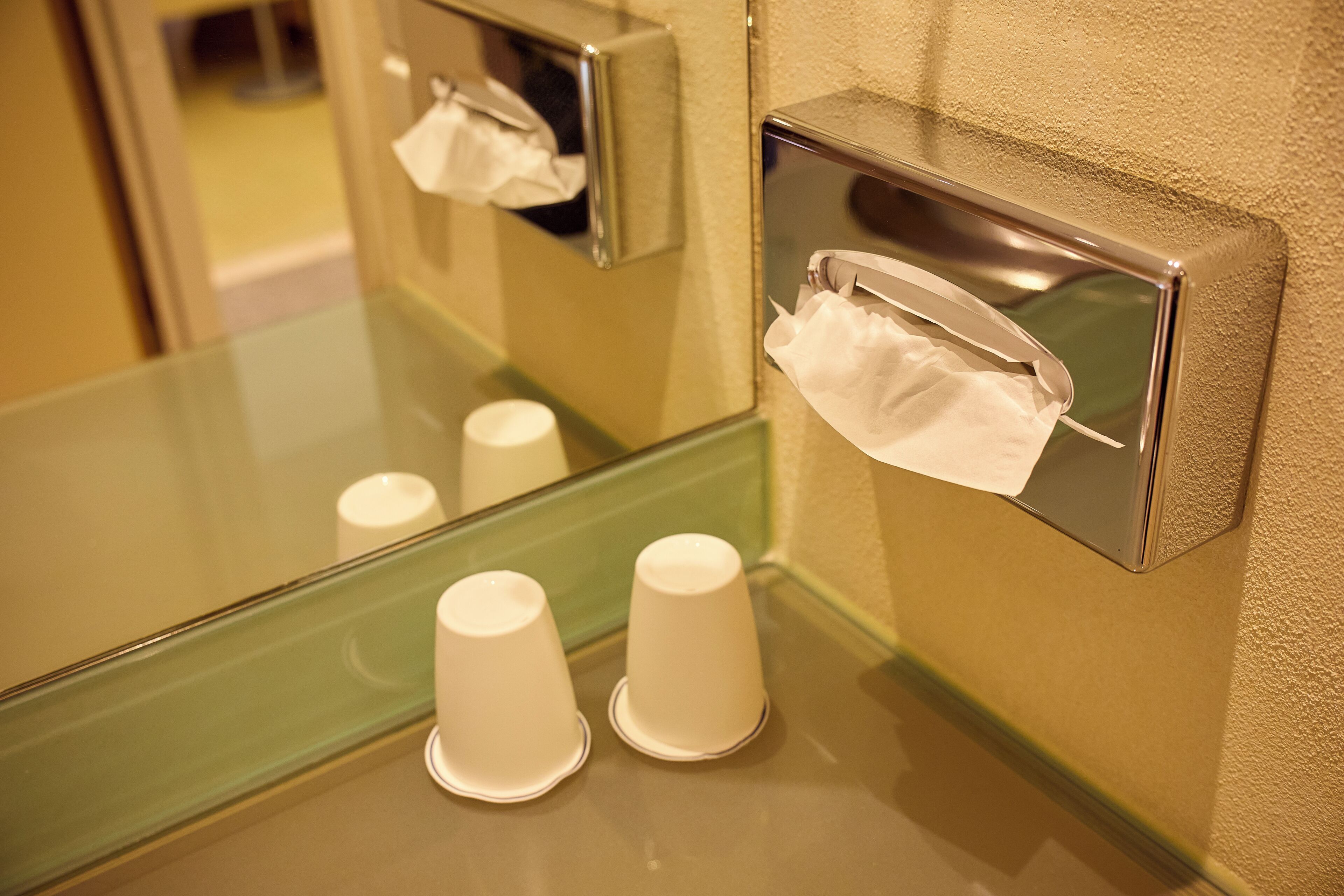A restroom with a mirror, a tissue dispenser, and two paper cups on the counter.