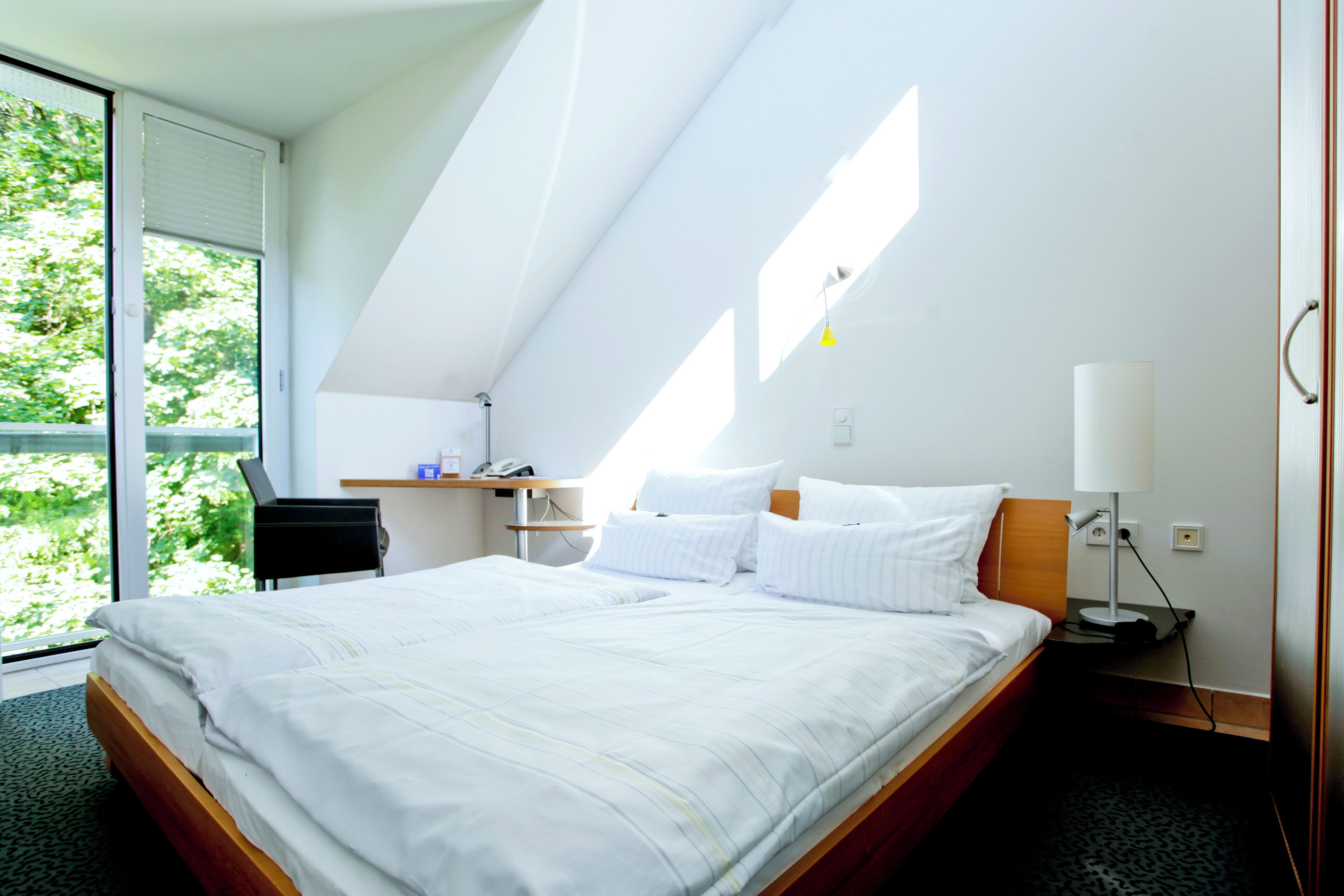 A hotel room with a bed, a desk, a chair, a lamp, and a window with a view of greenery.