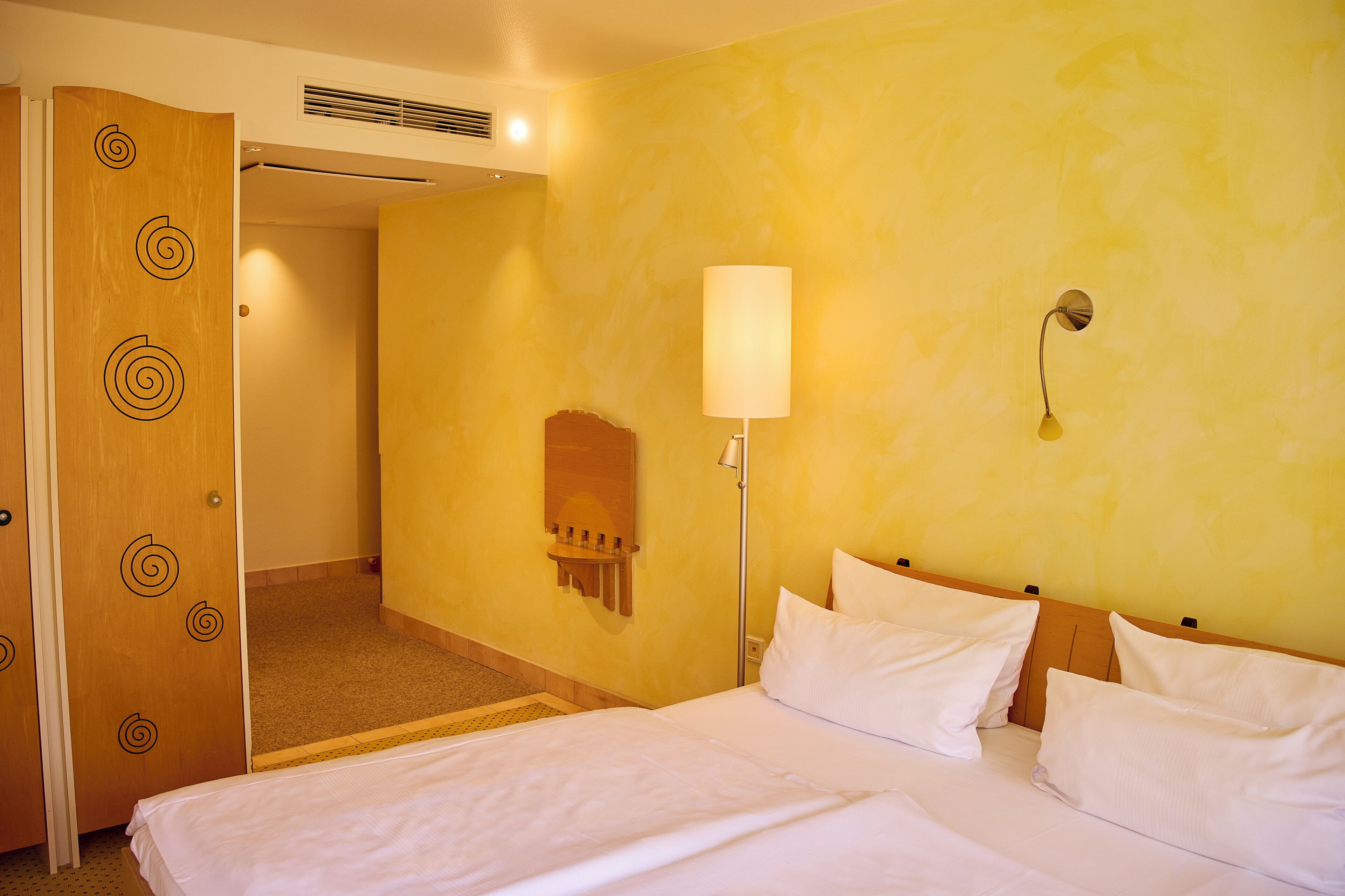 Quarto de hotel com cama, luminária fixada na parede, porta de madeira com desenhos espirais e uma pequena prateleira.