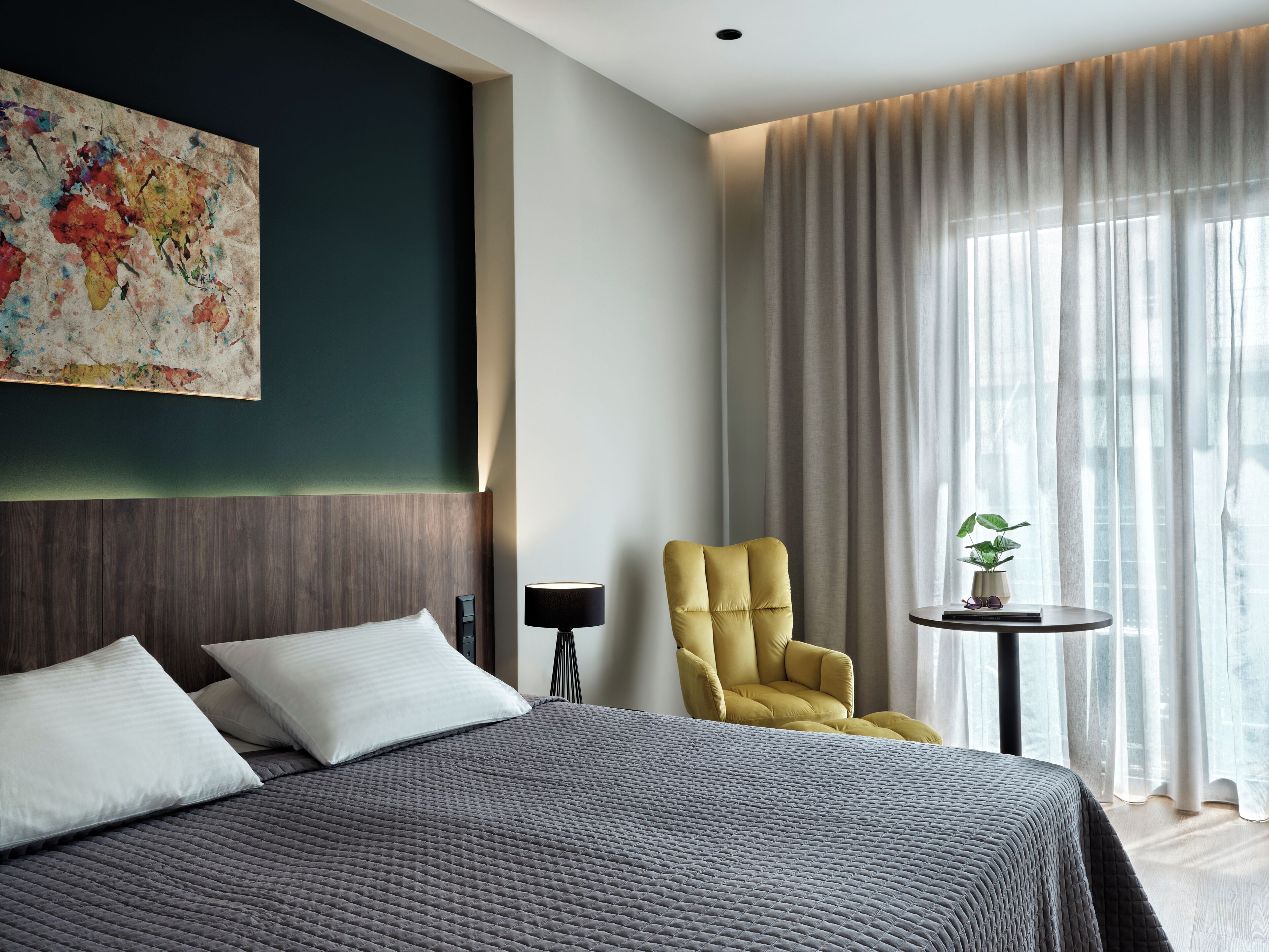A modern hotel room with a large bed, a yellow armchair, a small table with a plant, and a large window with sheer curtains.