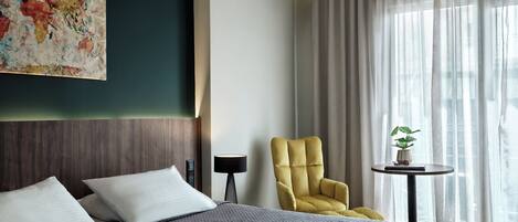 A modern hotel room with a large bed, a yellow armchair, a small table with a plant, and a large window with sheer curtains.