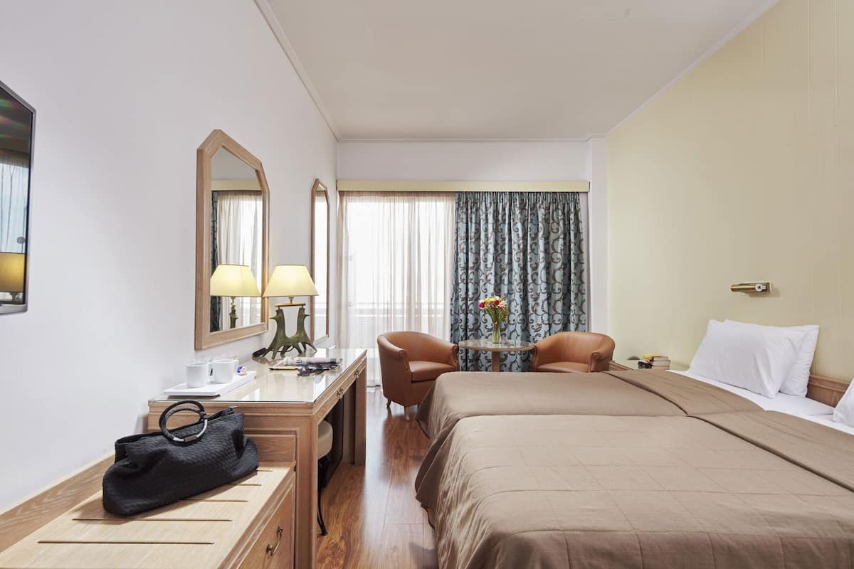 A hotel room with a bed, two chairs, a desk with a lamp, a mirror, and a window with curtains.