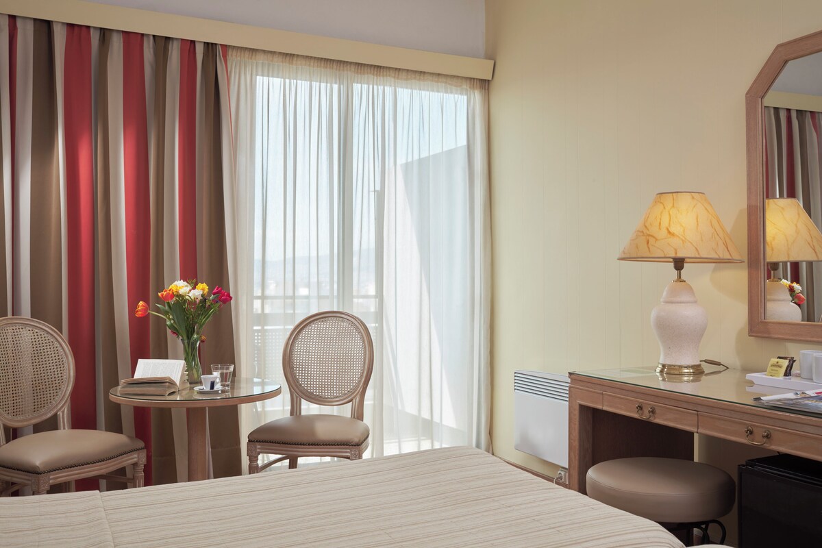 A hotel room with a bed, a desk with a lamp, a chair, a small table with a vase of flowers, and a window with curtains.