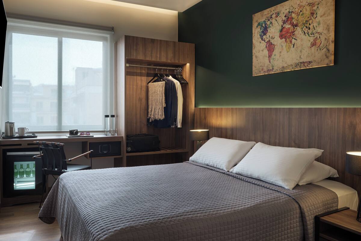 A modern hotel room with a large bed, a desk with a computer, a wardrobe, and a map on the wall.