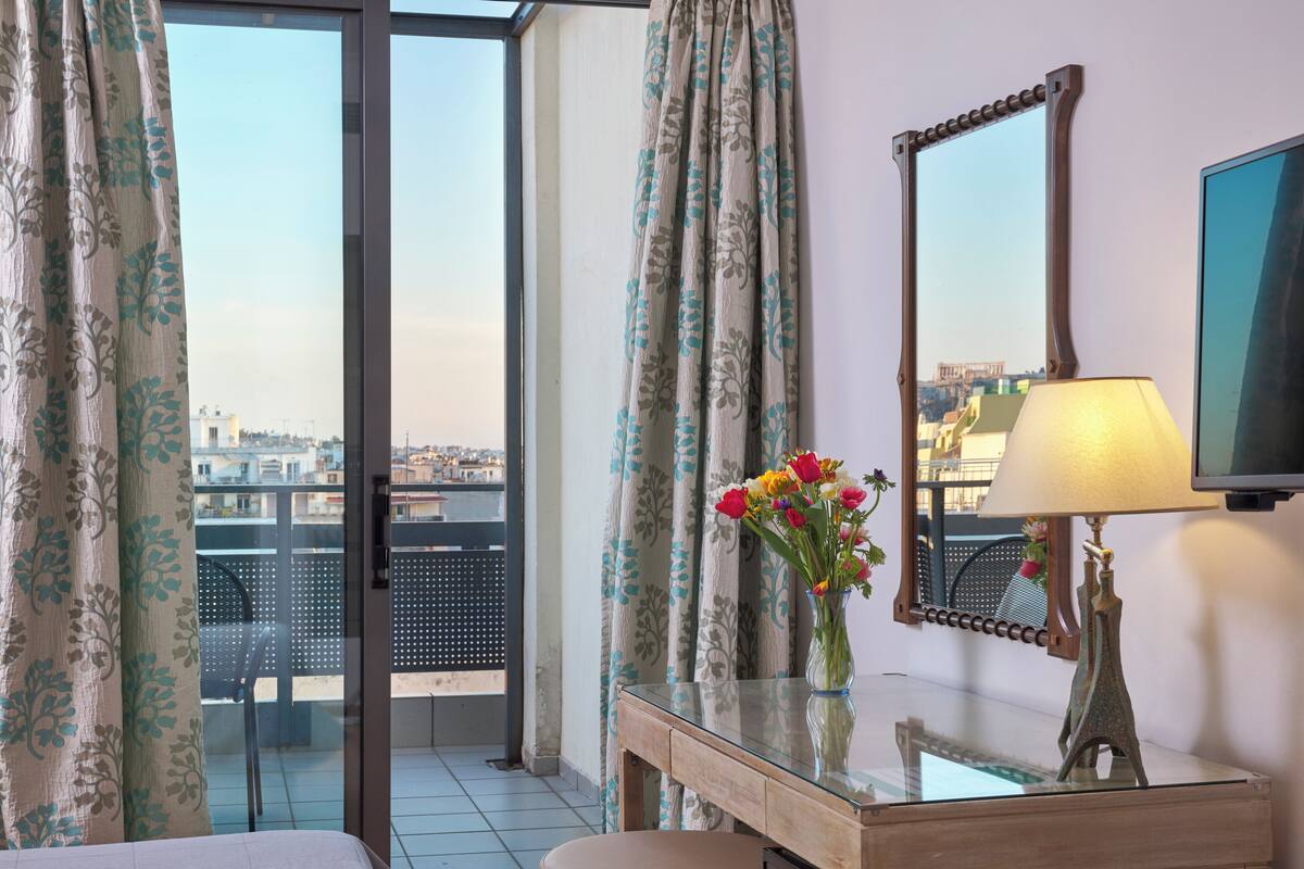 A hotel room with a balcony, a desk with a lamp, a vase of flowers, and a large mirror.