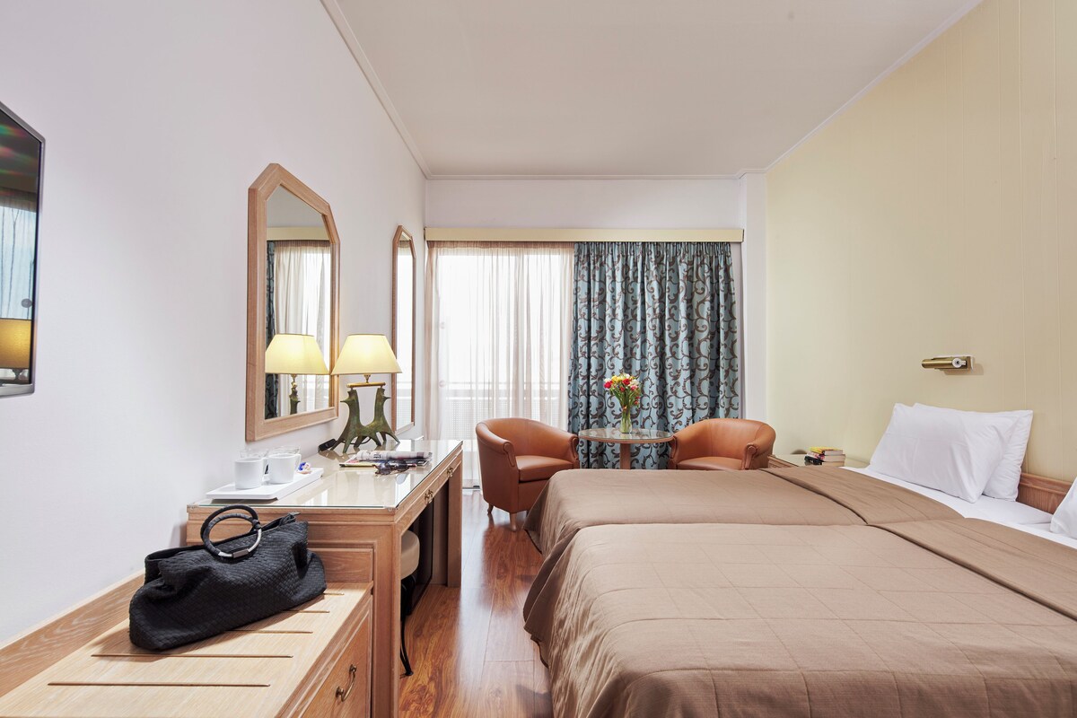 A hotel room with a bed, two chairs, a desk with a lamp, a mirror, and a handbag on the desk.