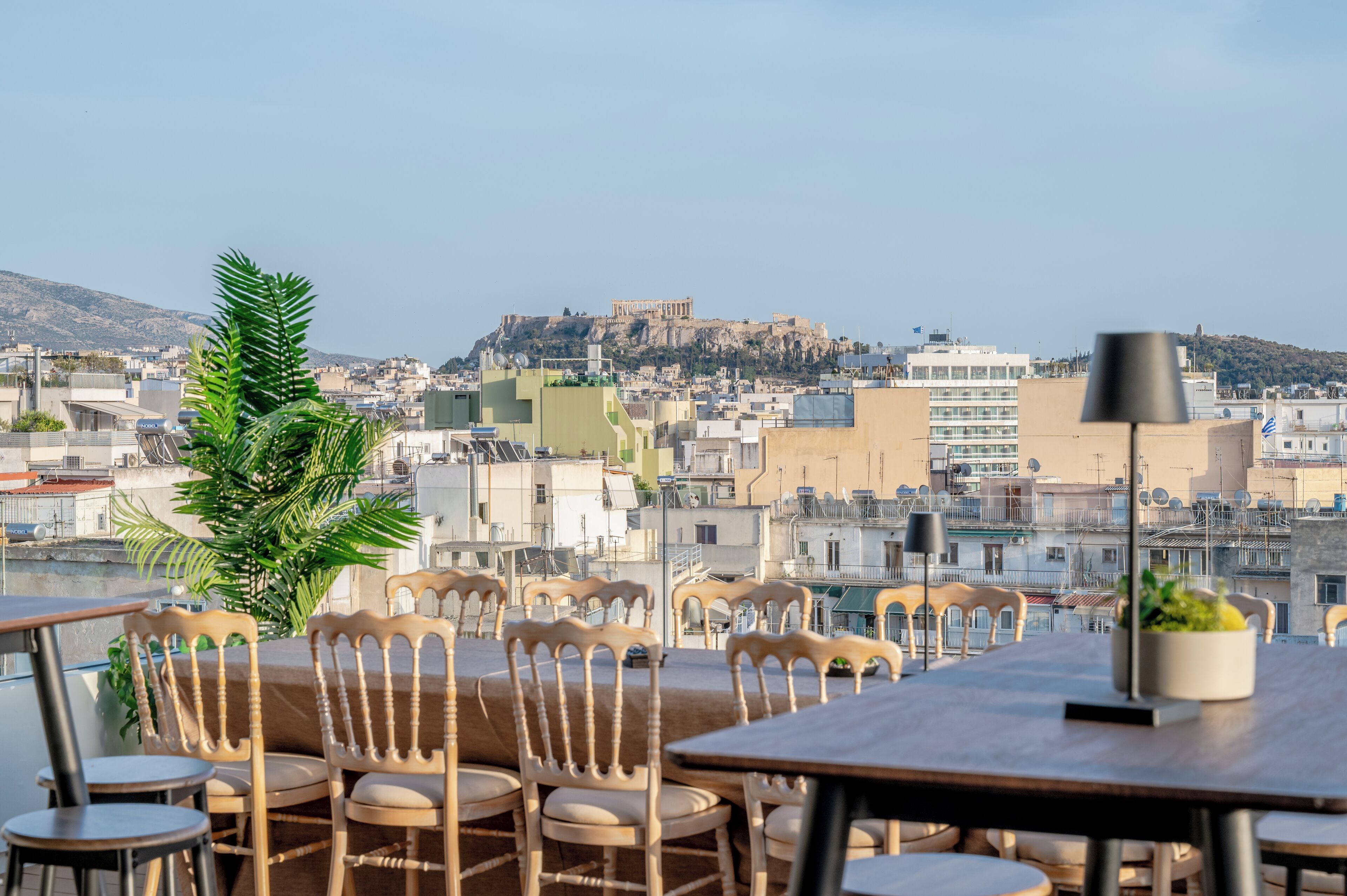 A rooftop terrace with wooden chairs and tables, a potted plant, and a view of a historic hilltop fortress.