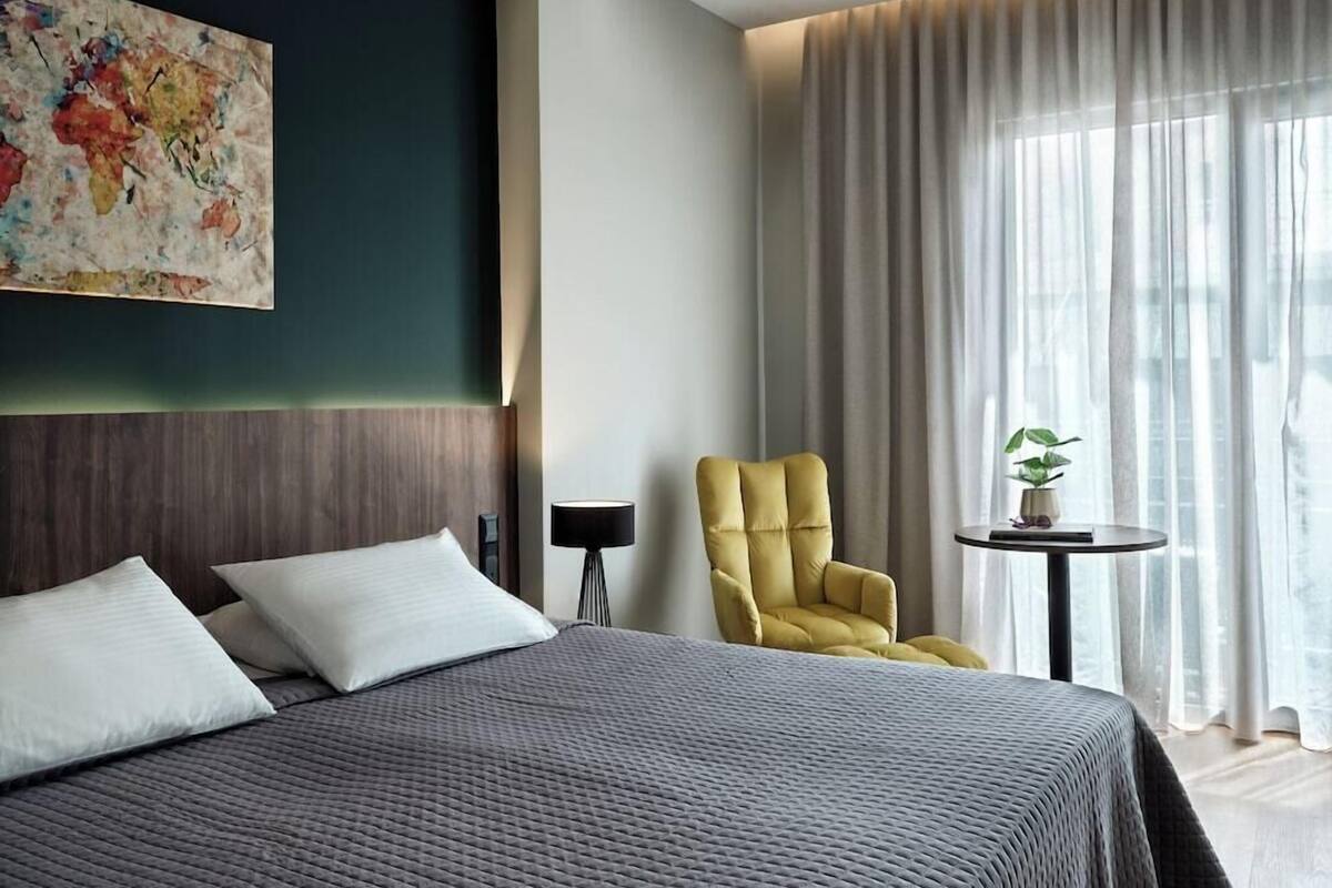 A modern hotel room with a large bed, a yellow armchair, a small table with a plant, and a large window with curtains.