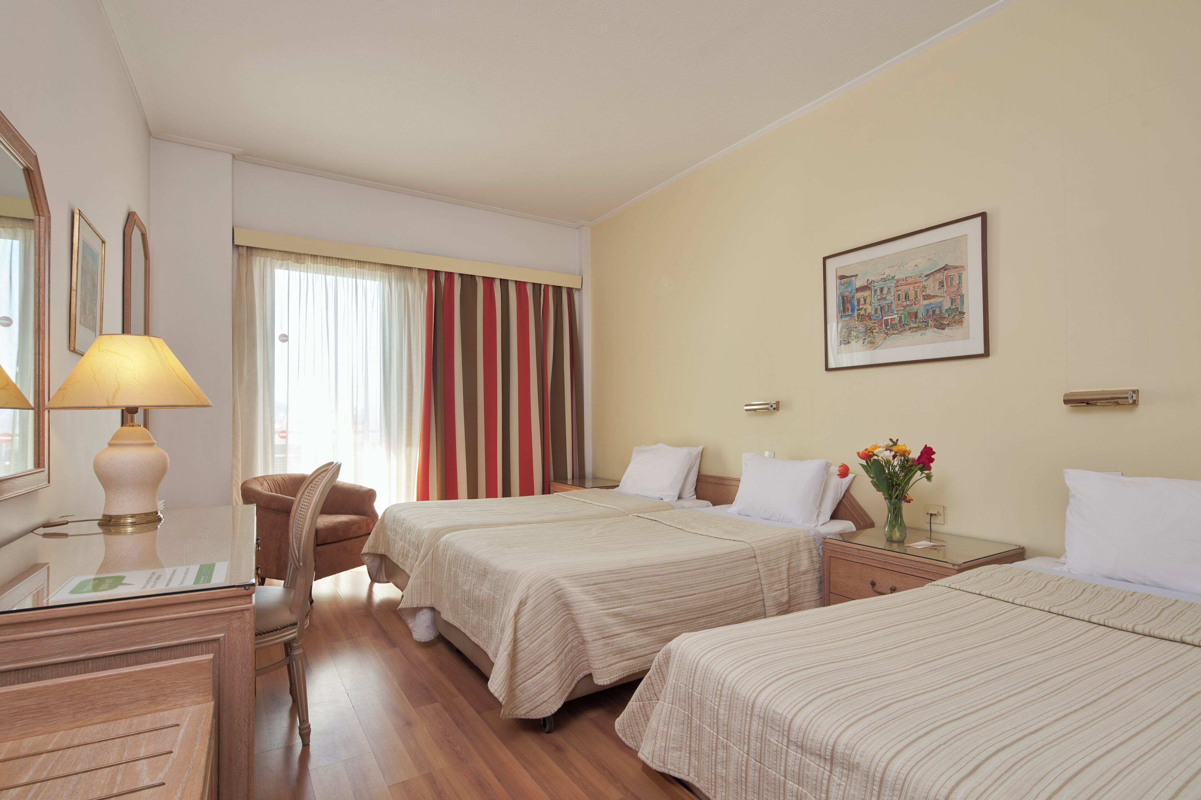 A hotel room with two beds, a desk with a lamp, a chair, a nightstand with a vase of flowers, and a painting on the wall.