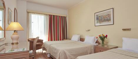 A hotel room with two beds, a desk with a lamp, a chair, a nightstand with a vase of flowers, and a painting on the wall.