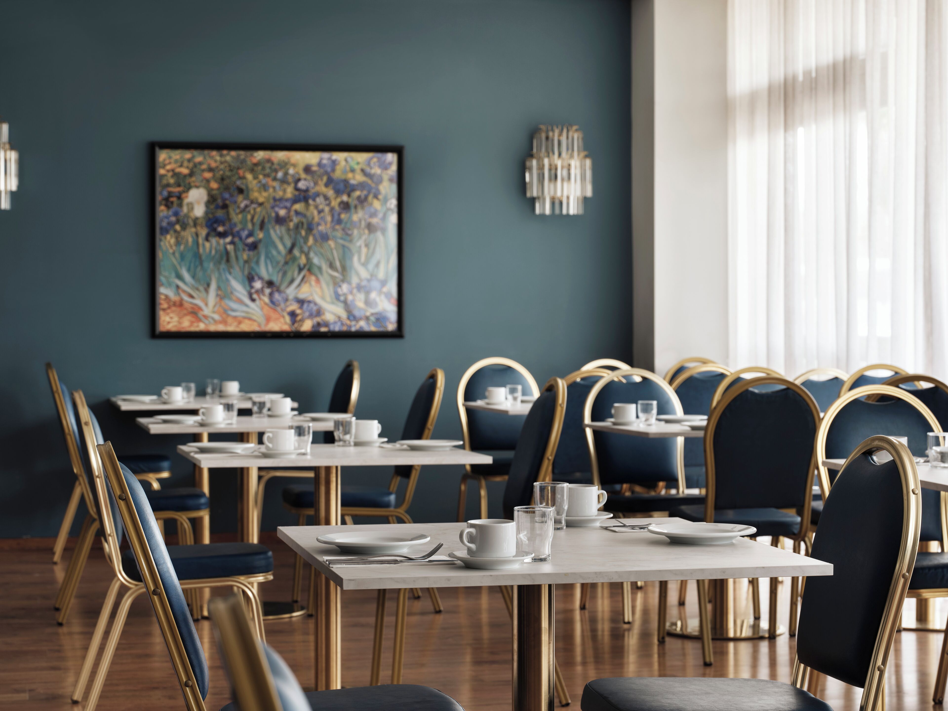 A restaurant with tables set for dining, chairs with gold frames, and a large floral painting on the wall.