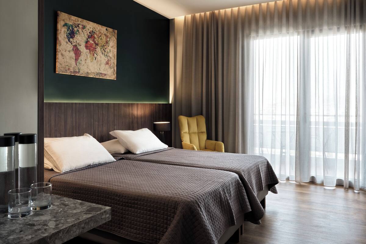 A hotel room with two beds, a large window with curtains, a modern lamp, and a painting on the wall.
