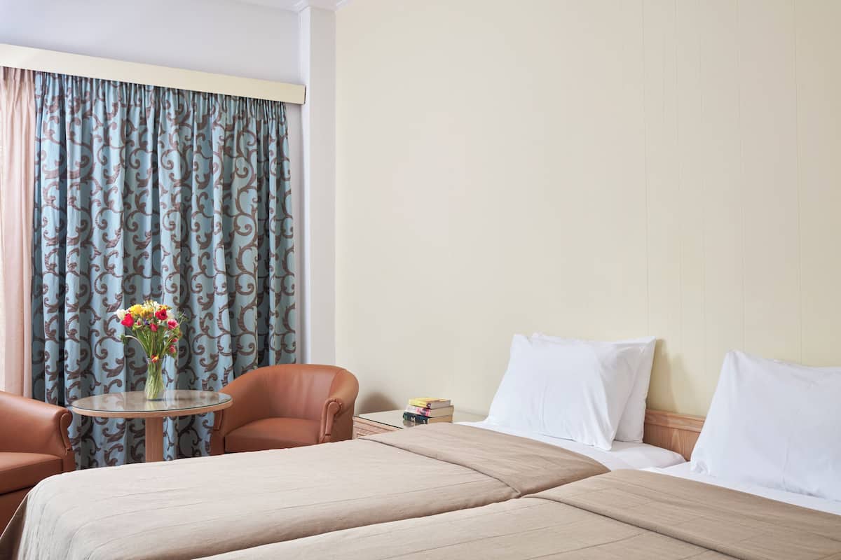 A hotel room with a bed, two armchairs, a small table with a vase of flowers, and a nightstand with books.