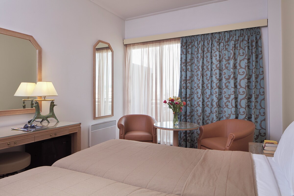 A hotel room with a bed, two chairs, a desk with a lamp, a mirror, and a window with curtains.
