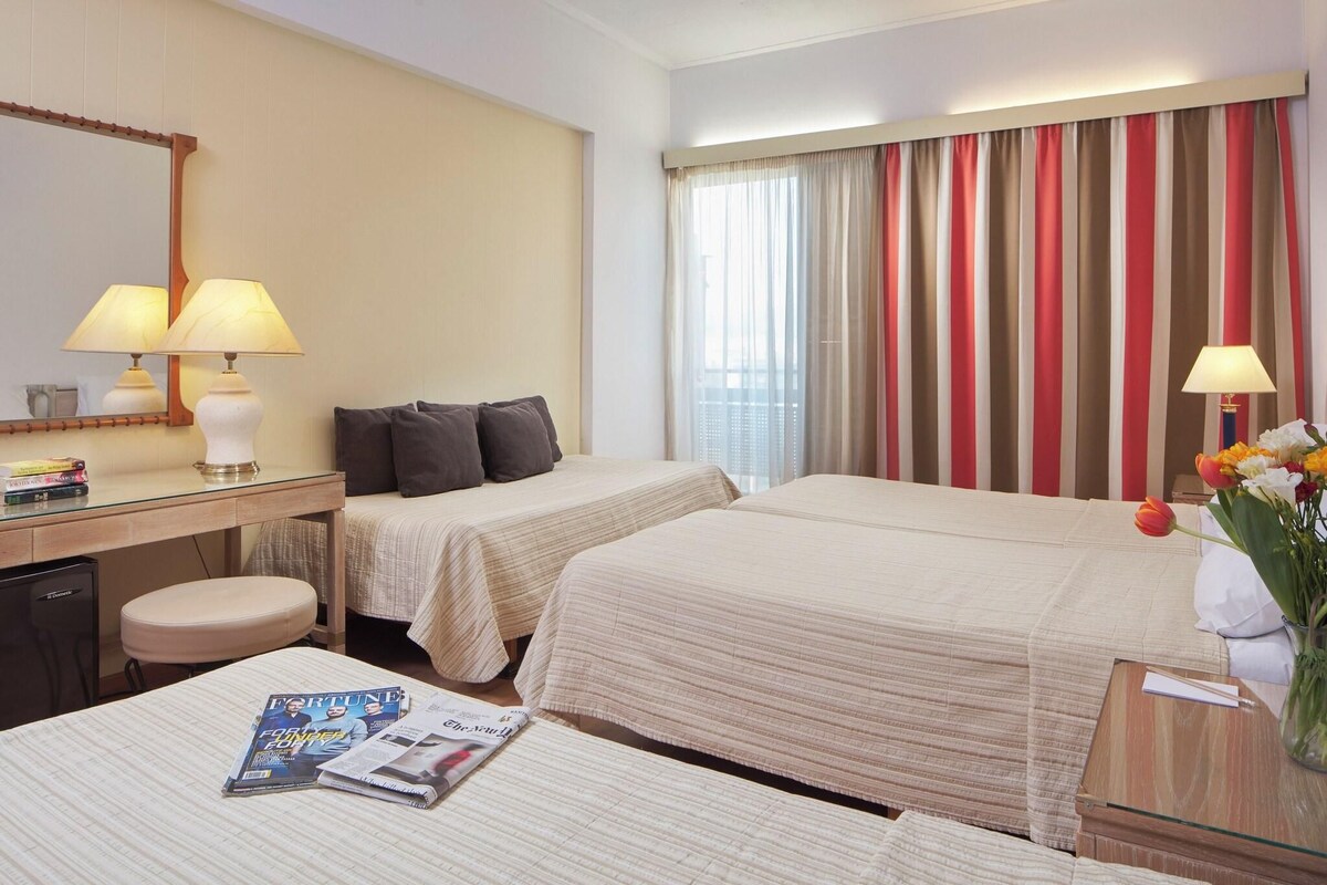 A hotel room with two beds, a desk with a lamp, a chair, a small table with a vase of flowers, and a window with striped curtains.