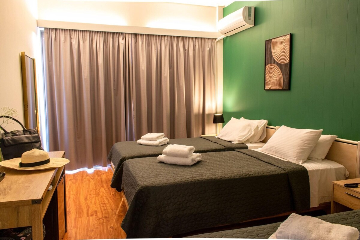 A hotel room with two beds, a desk, a chair, a hat, and a wall-mounted air conditioner.