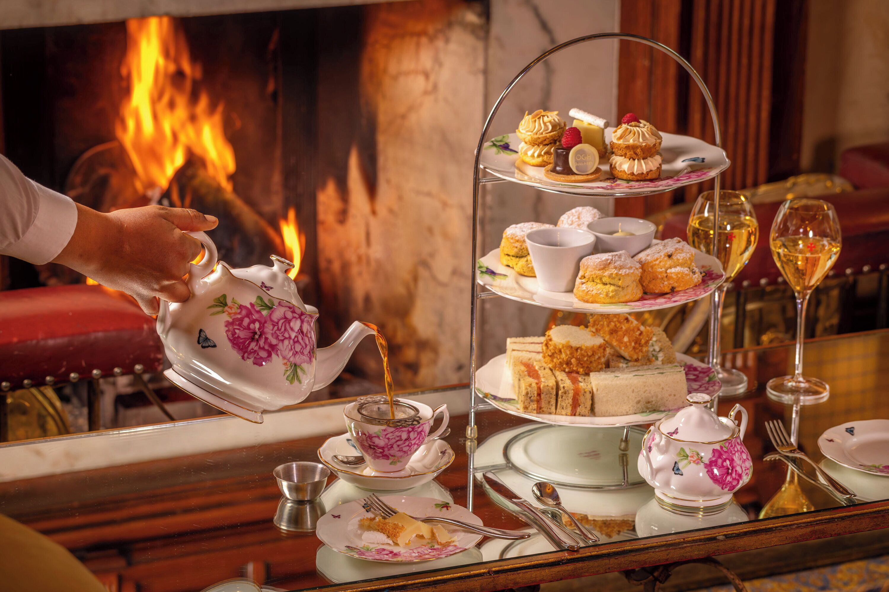 Een theeservies met een theepot die thee inschenkt, een etagère met desserts en een glas wijn op een reflecterend oppervlak.