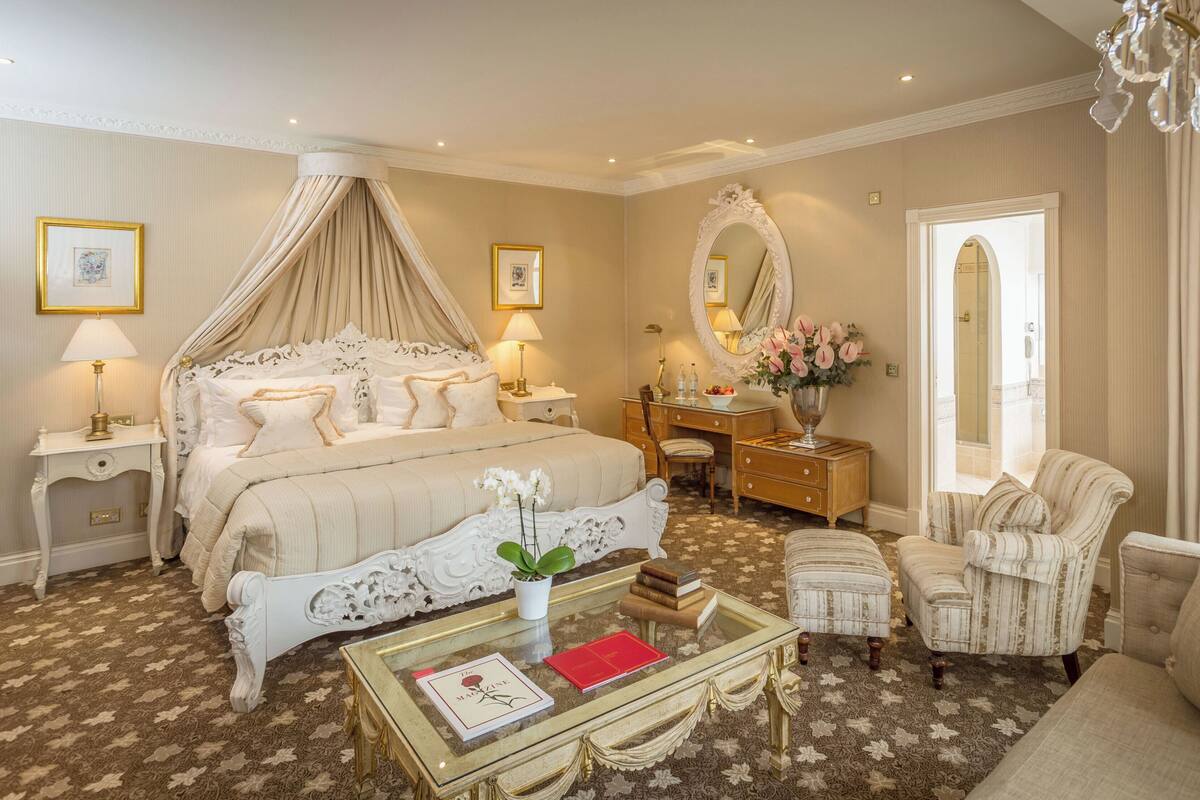 A luxurious bedroom with a canopy bed, a vanity with a mirror, a chaise lounge, and a glass coffee table with books.
