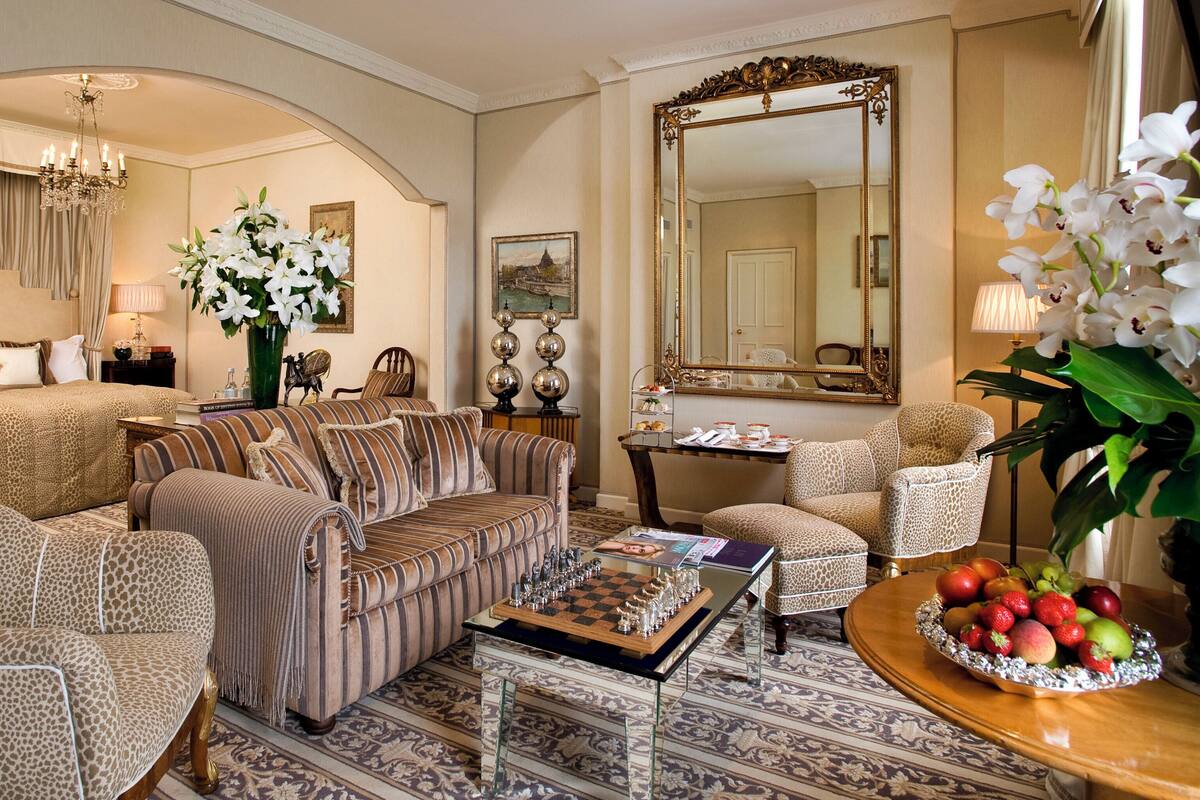 A well-appointed hotel room with a large bed, a sofa, a coffee table with a chess set, a mirror, and a fruit bowl.