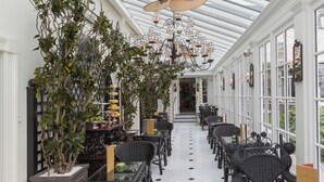A conservatory with a glass ceiling, wicker furniture, potted plants, and a chandelier.