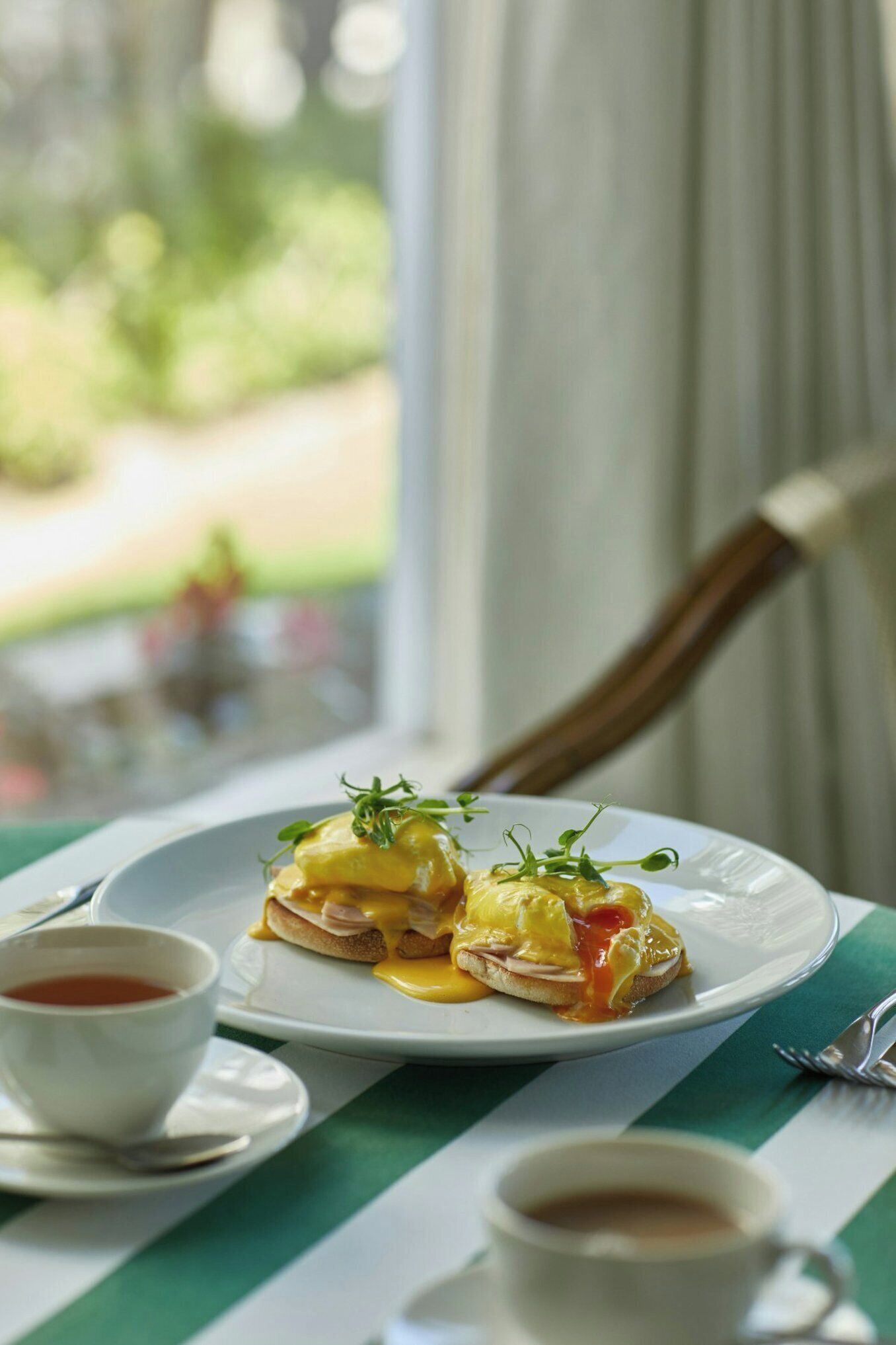 A breakfast setting with eggs Benedict on a plate, a cup of tea, and a cup of coffee on a table with a striped tablecloth.