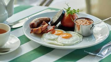 A breakfast setting with a plate of eggs, sausages, and bacon, a small cup of coffee, and a teapot on a striped tablecloth.