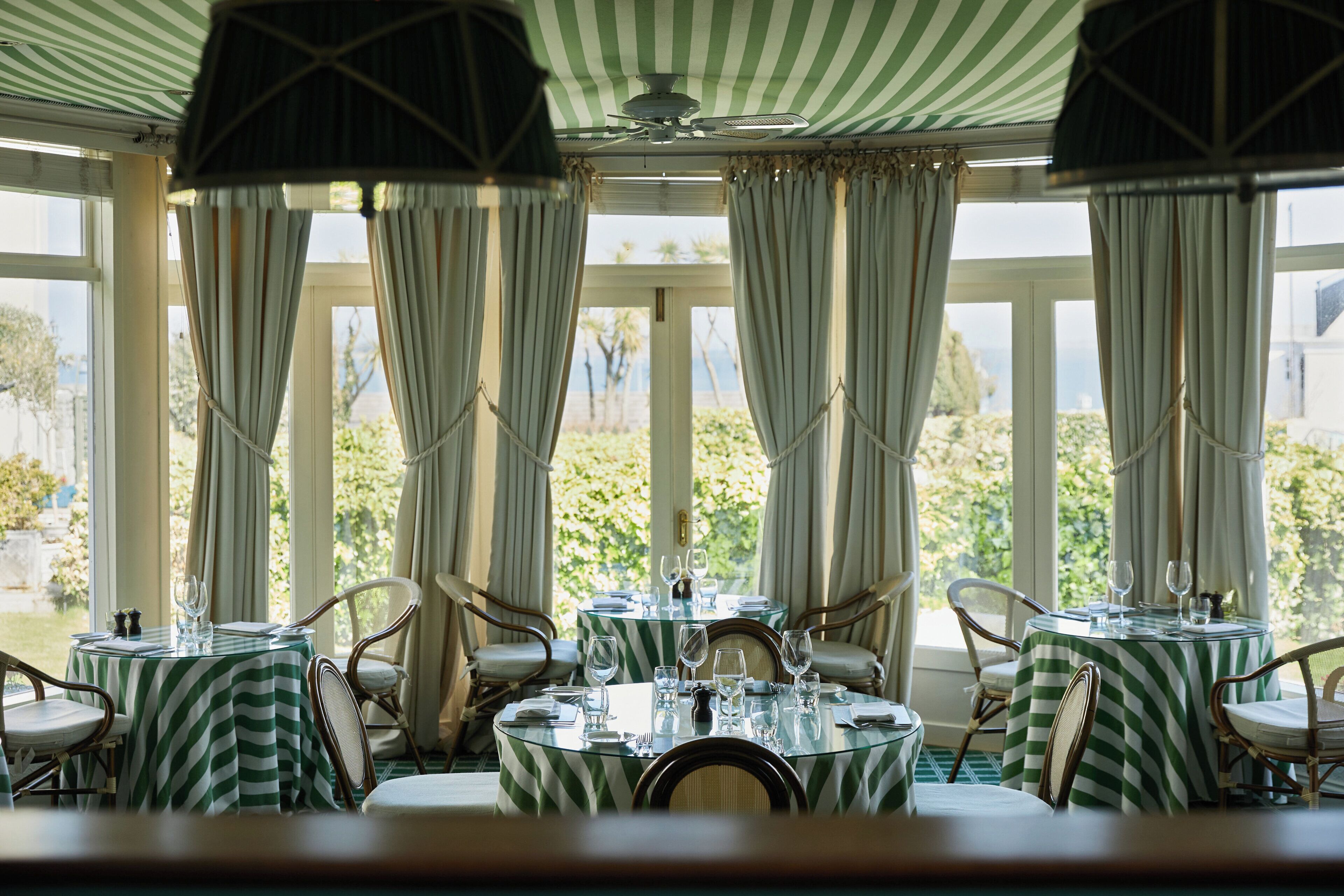 A dining area with round tables, chairs, and a view of greenery through large windows.