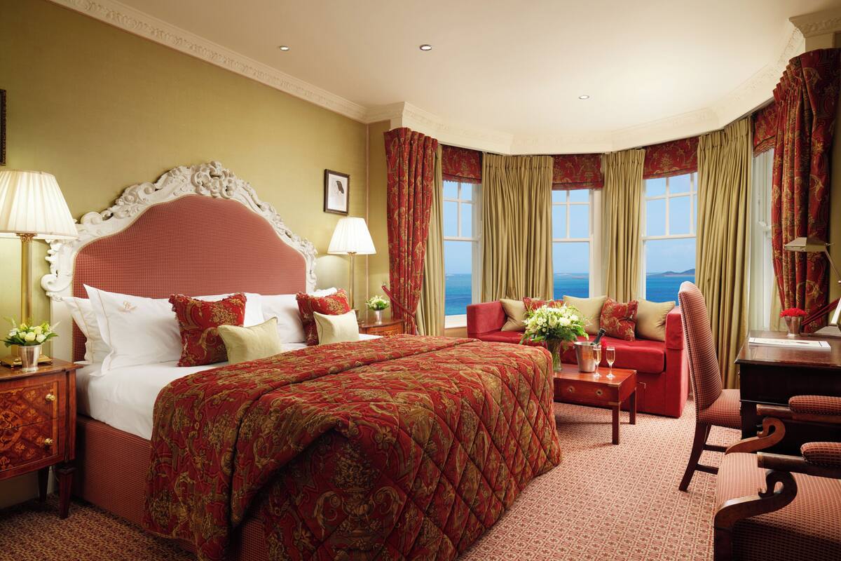 A hotel room with a large bed, a sitting area with a red sofa, a desk, and a view of the sea.