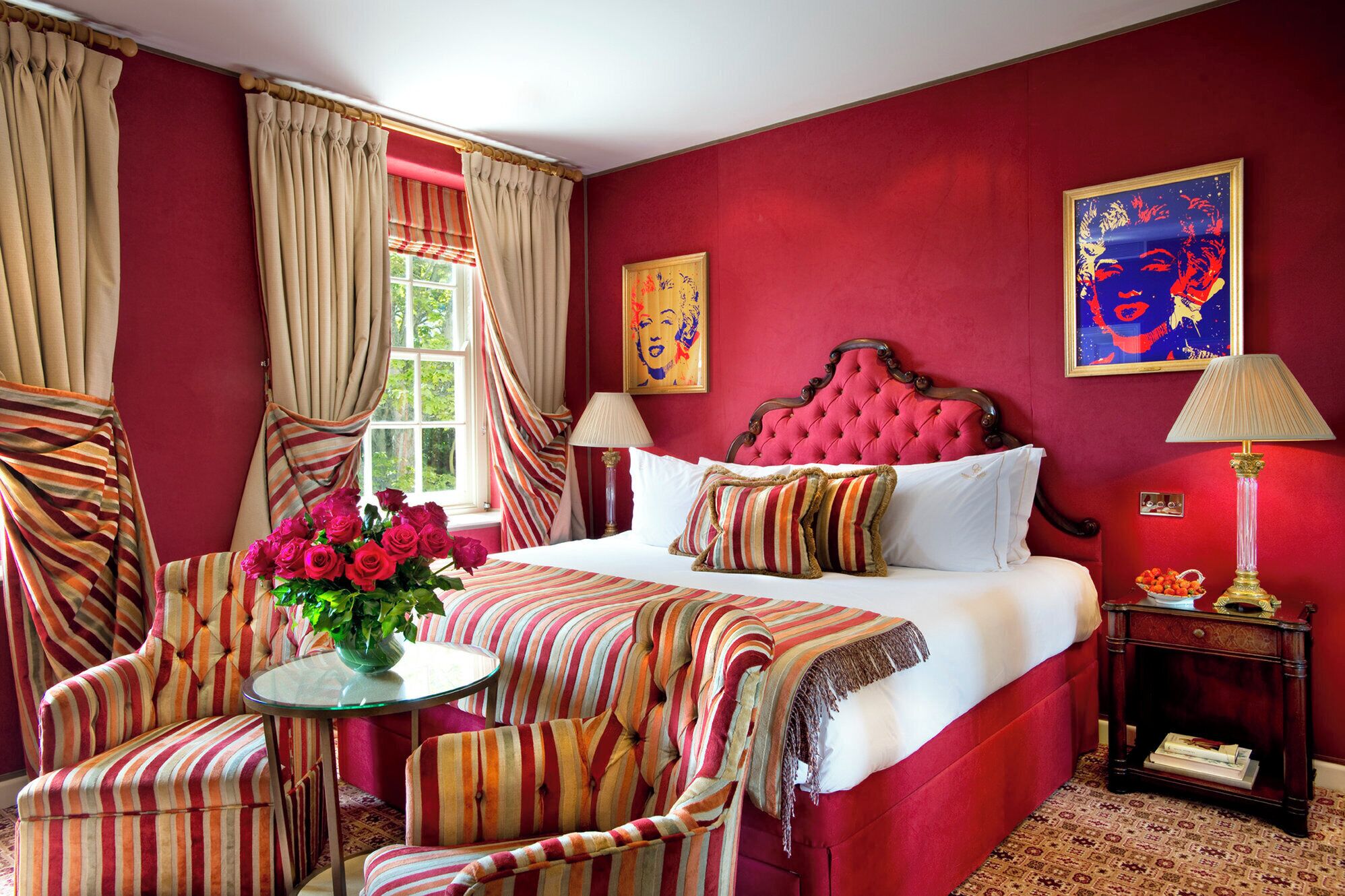 A hotel room with a red wall, a tufted headboard, a bed with striped bedding, a striped armchair, a small table with a vase of flowers, and two lamps.