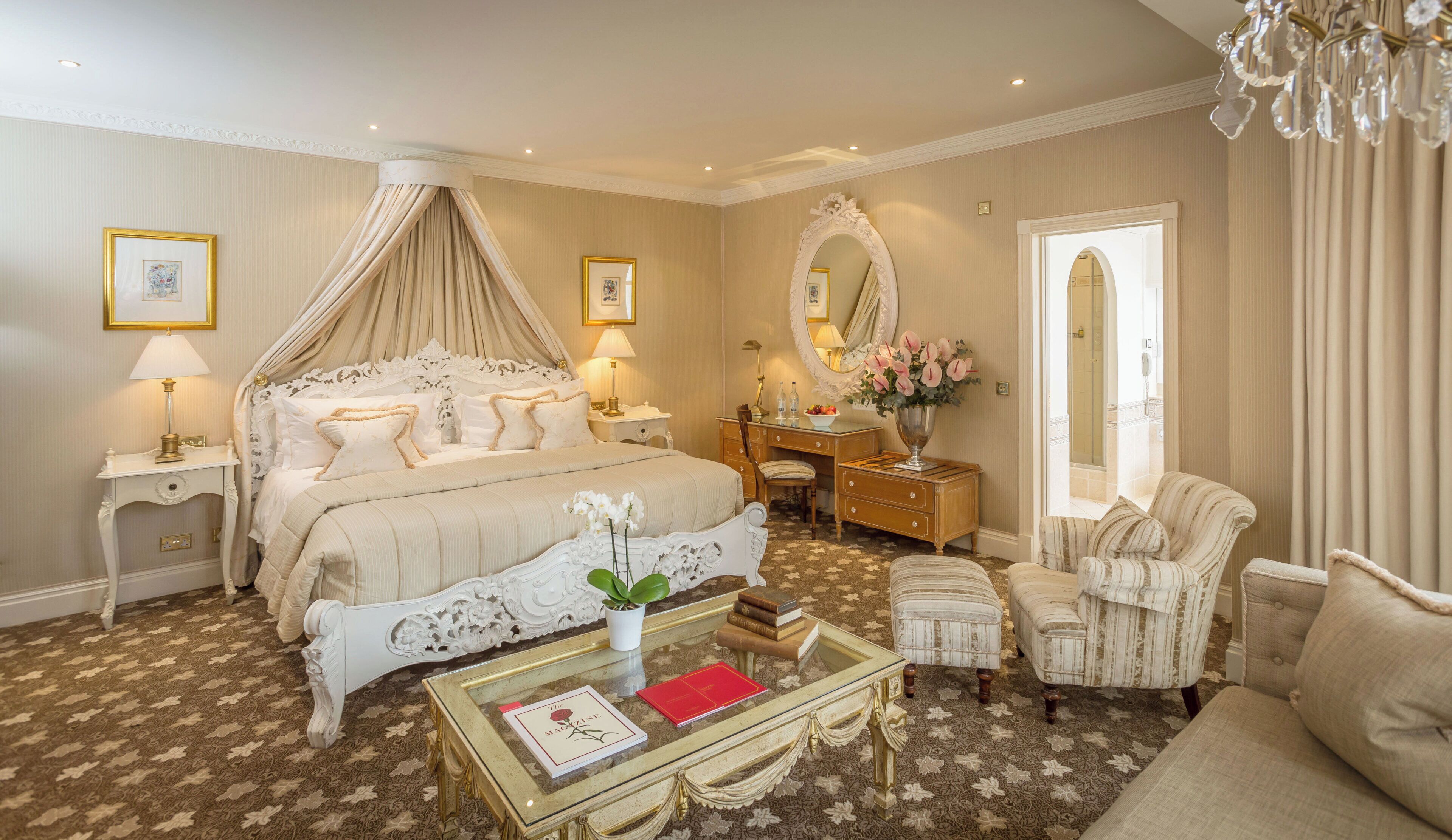A luxurious bedroom with a canopy bed, a vanity with a mirror, a chaise lounge, and a glass coffee table with books.