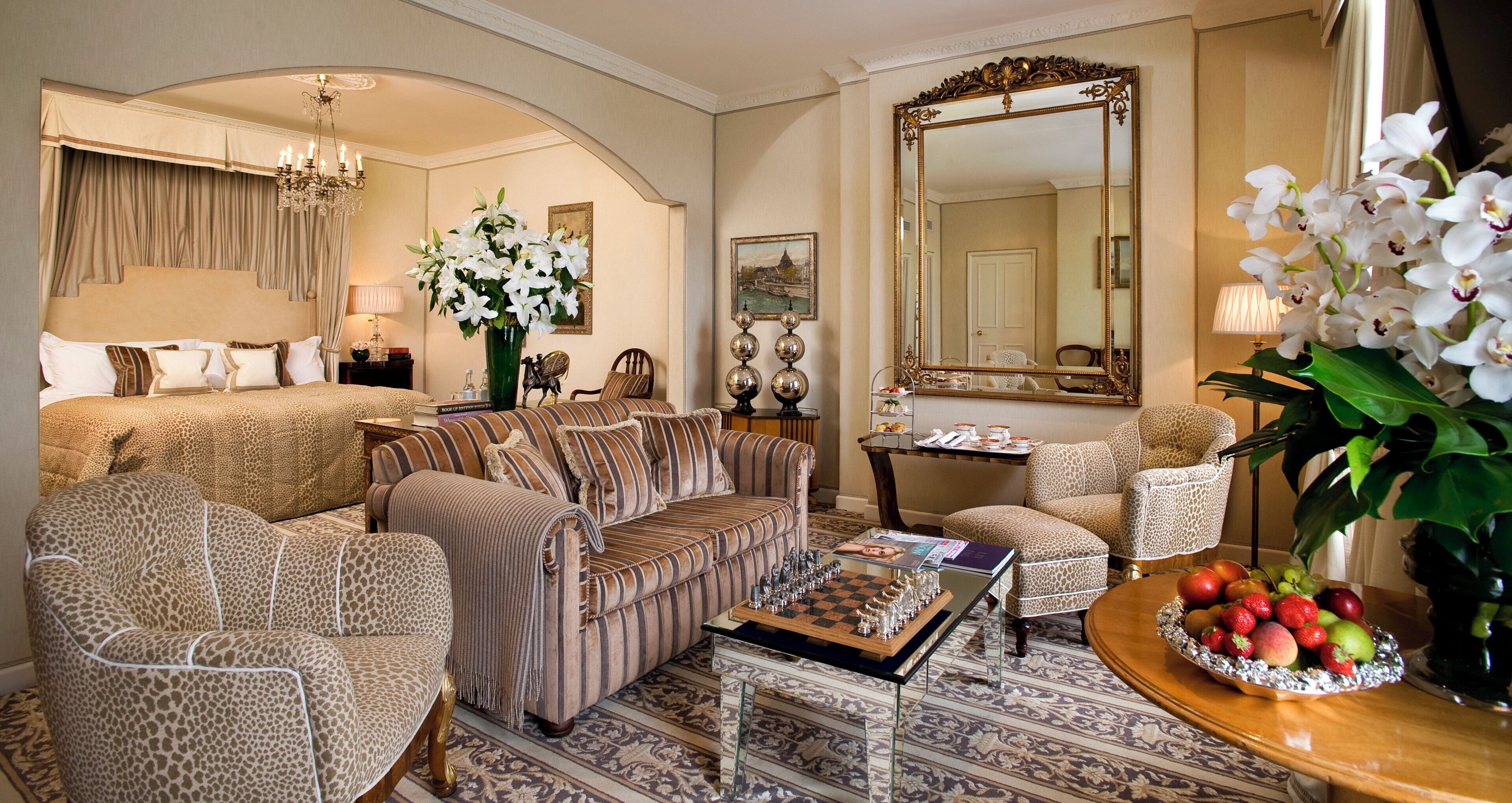 A well-appointed hotel room with a large bed, a sofa, a coffee table with a chess set, a mirror, and a fruit bowl.