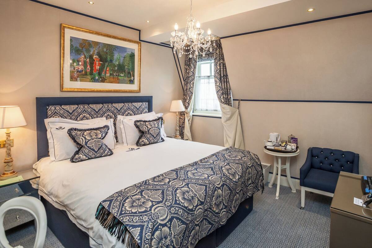 A hotel room with a large bed, a small table with a chair, a television, and a painting on the wall.