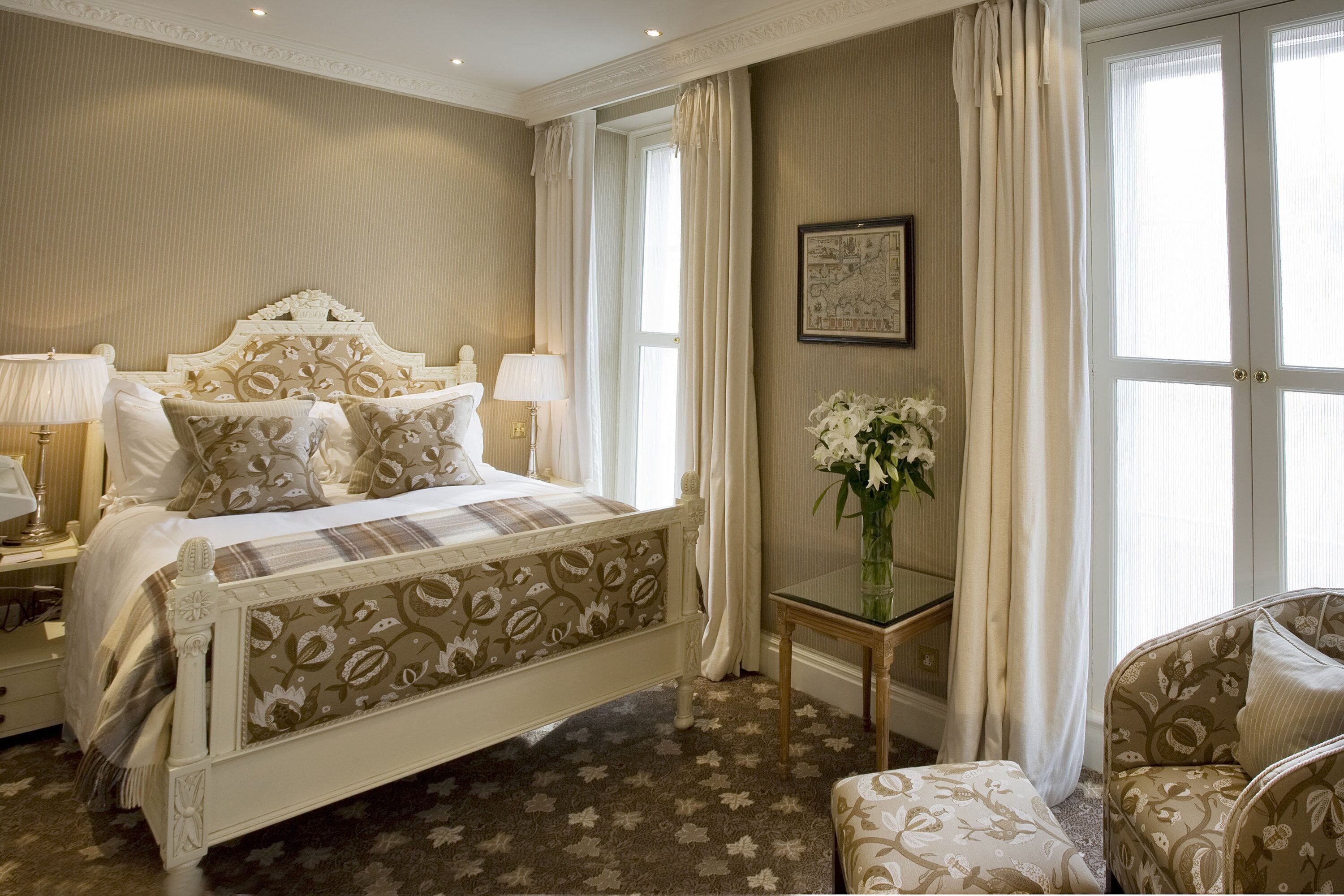 Een slaapkamer met een bed, nachtkastjes, een stoel, een klein tafeltje en een raam met gordijnen.