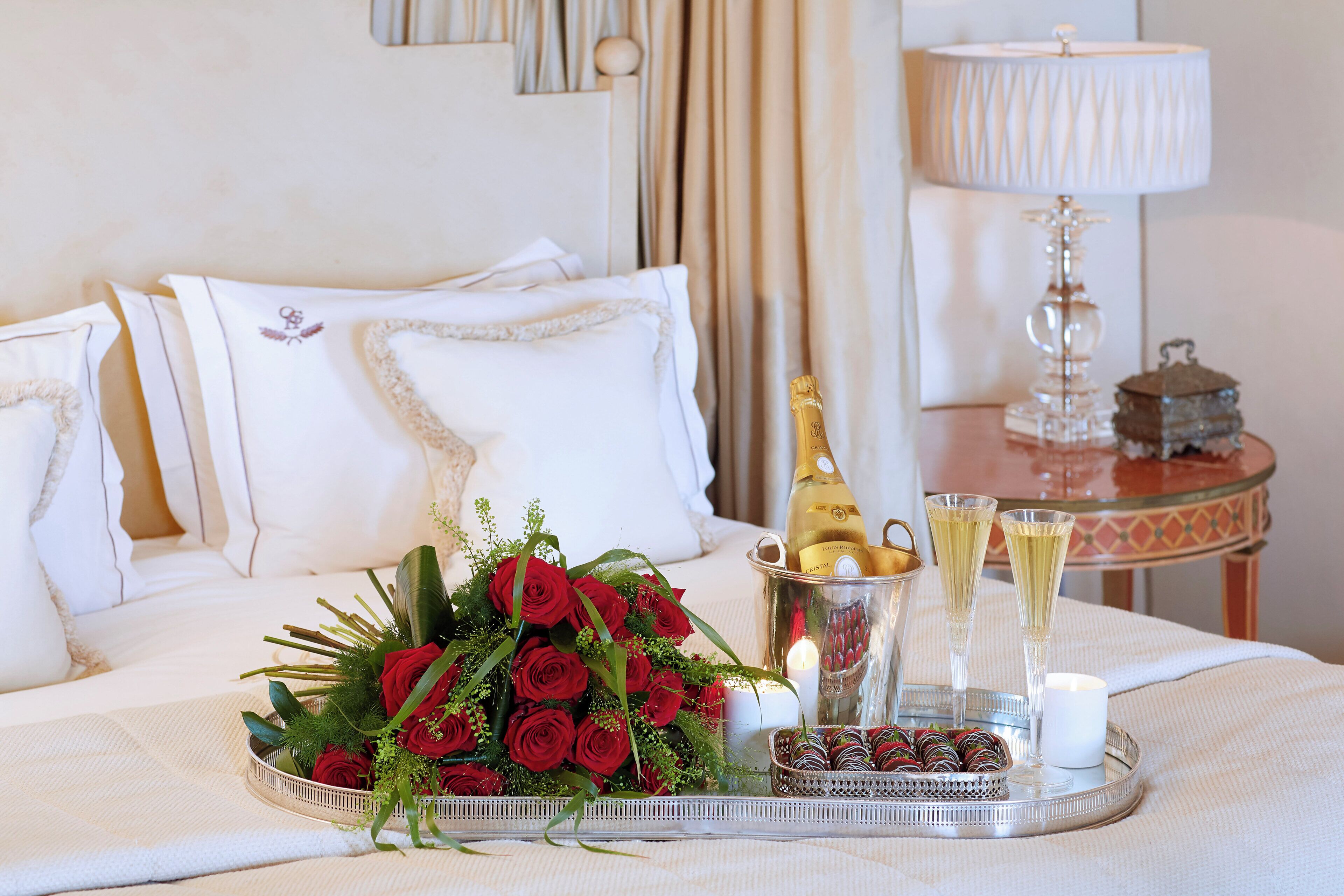 A bed with white linens, a bouquet of red roses, a champagne bottle, and two glasses of champagne on a tray.