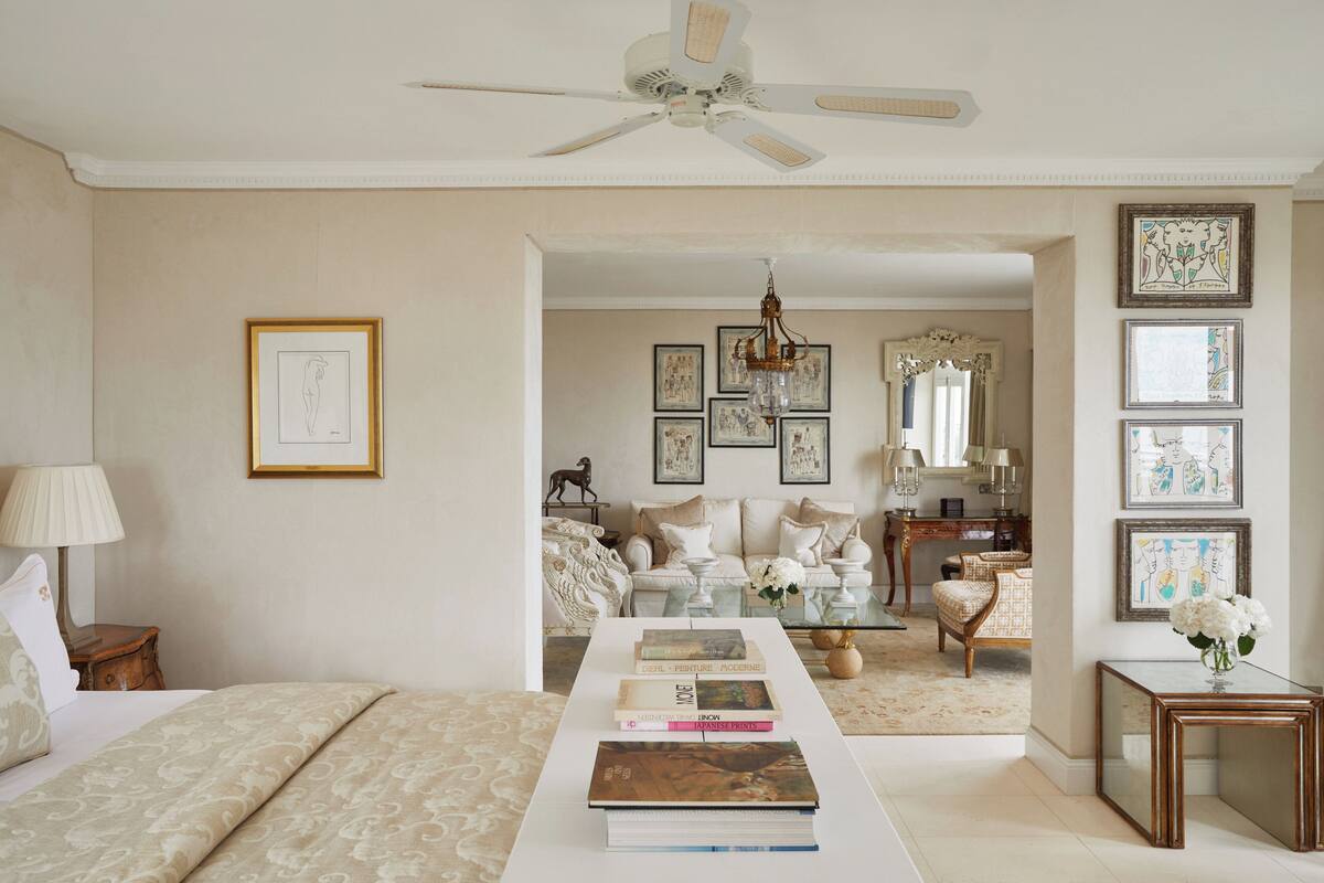 A bedroom with a bed, a bedside table, a nightstand, a ceiling fan, a coffee table with books, a mirror, and a door leading to a living area.