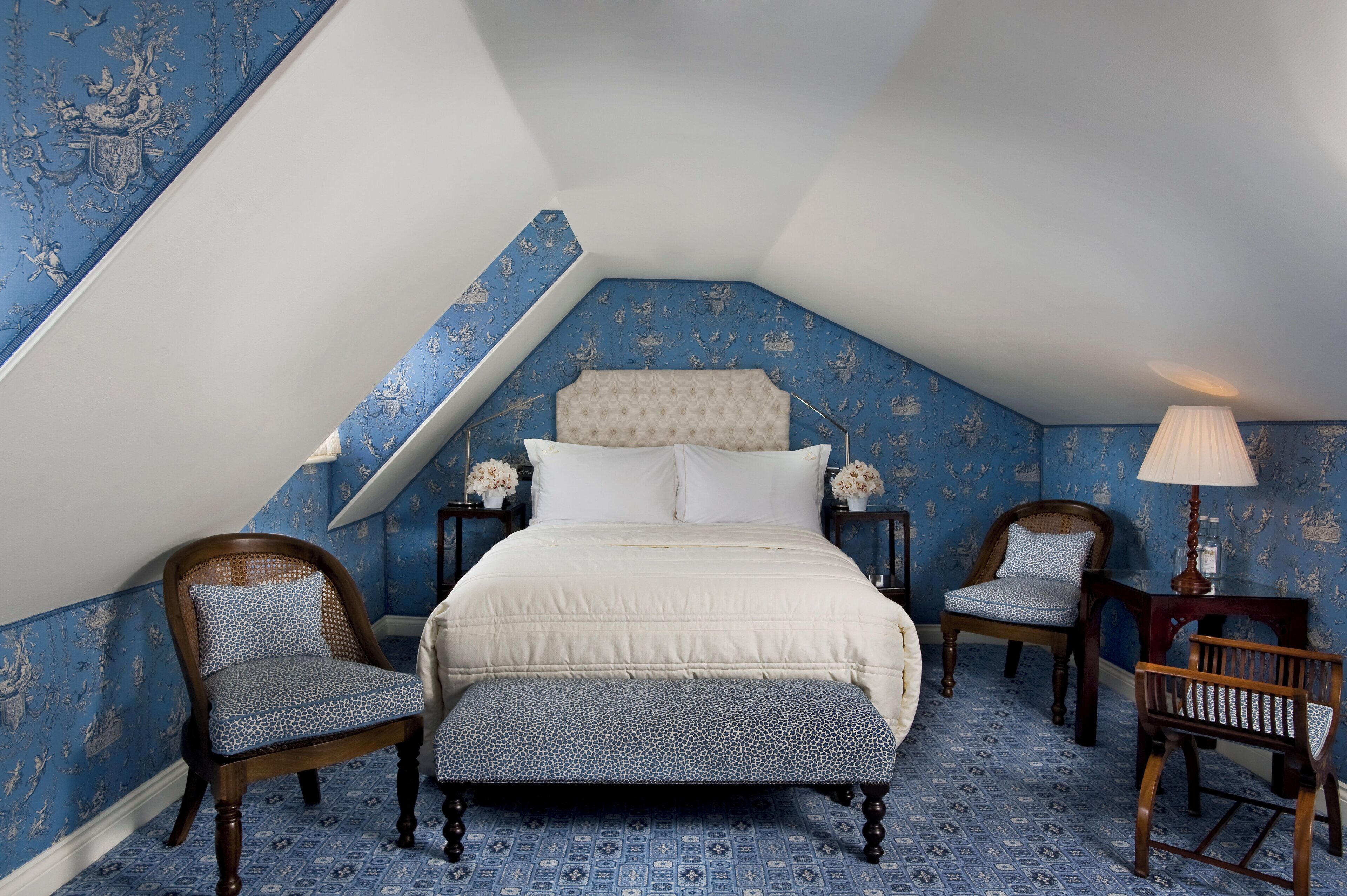 Ein Schlafzimmer mit einem Bett, zwei Stühlen, einer Bank und einem Nachttisch mit einer Lampe.