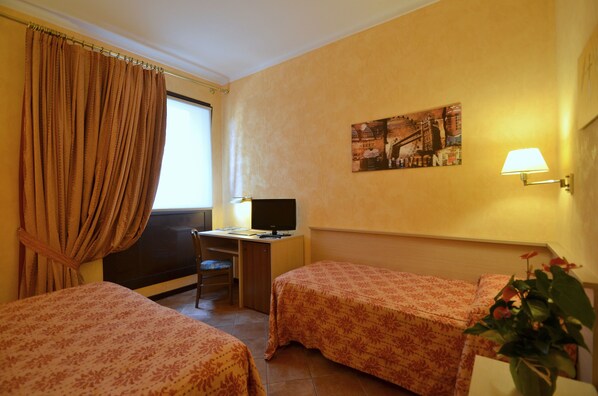 Une chambre d’hôtel avec deux lits, un bureau avec un ordinateur, une fenêtre avec des rideaux, une lampe fixée au mur et un tableau.