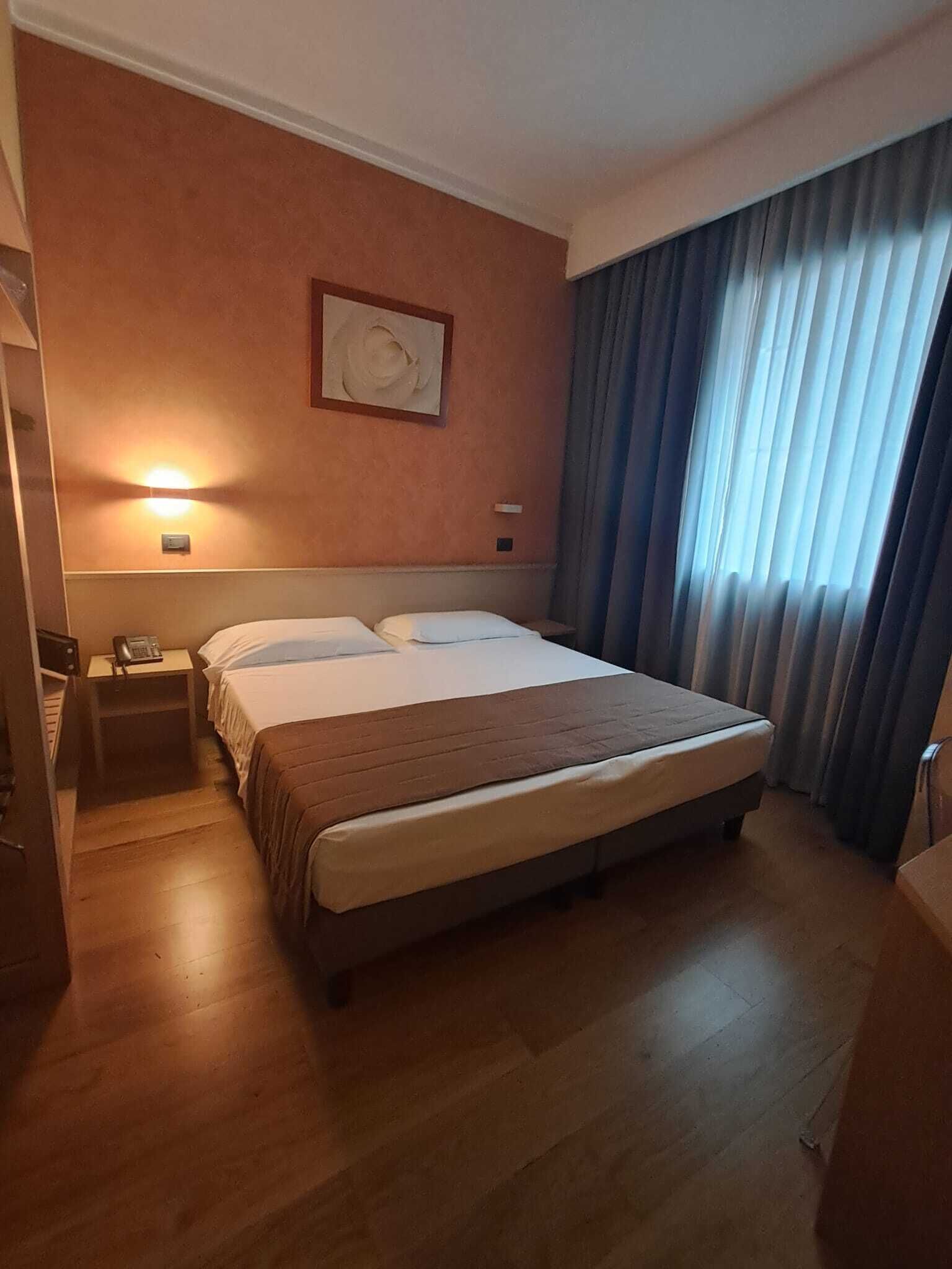 Ein Hotelzimmer mit einem großen Bett, einem Nachttisch mit Lampe, einem Wandbild und einem Fenster mit Vorhängen.