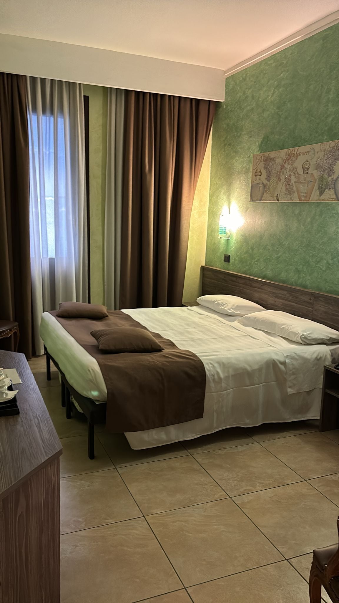 Una habitación de hotel con cama, cortinas, mesita de noche, escritorio y lámpara de pared.