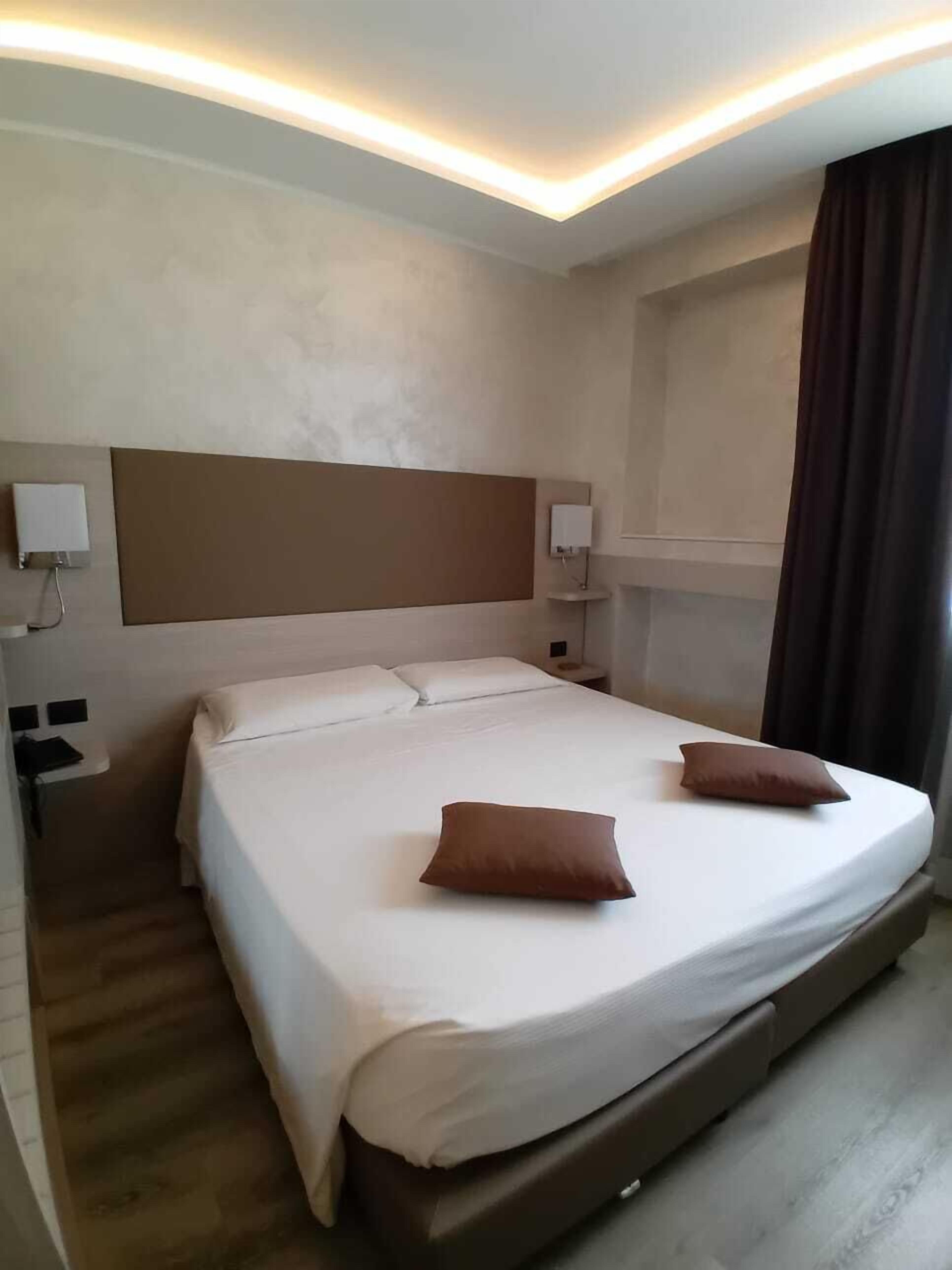 A hotel room with a large bed, two pillows, a nightstand, and a wall-mounted lamp.