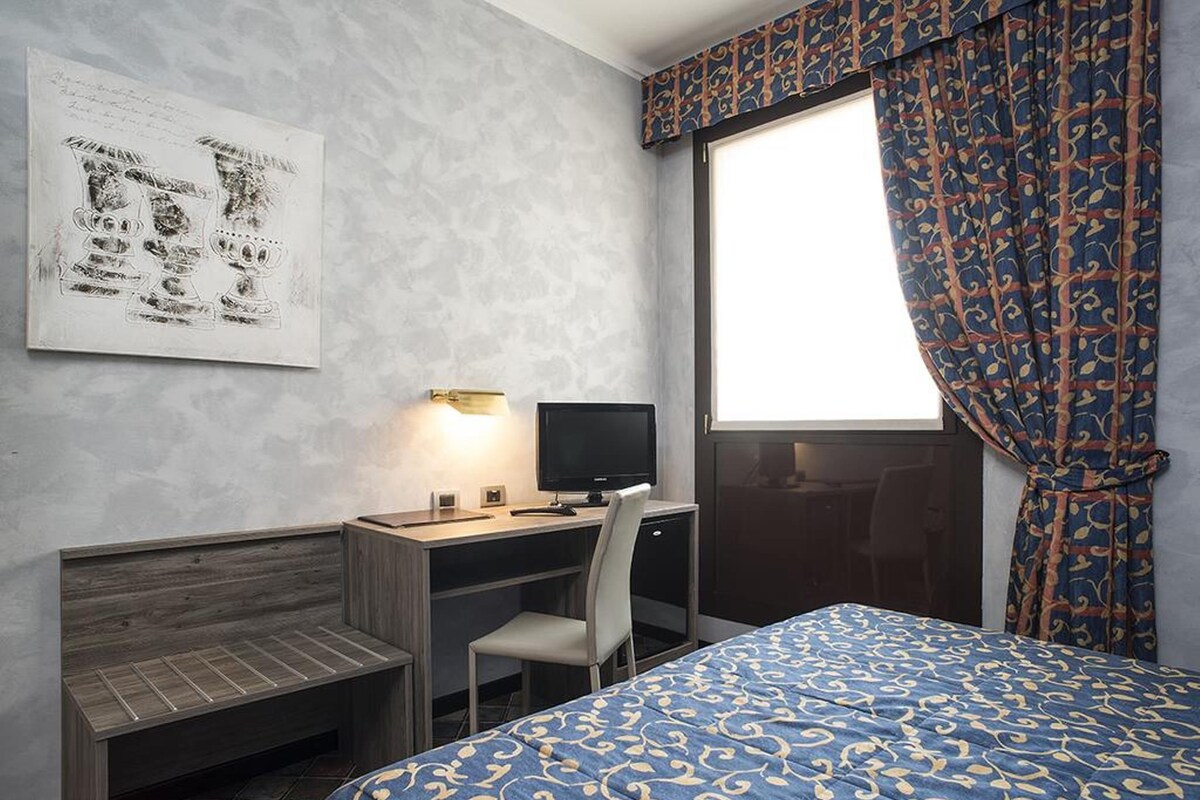 A hotel room with a bed, a desk with a computer, a chair, a window with curtains, and a wall with a framed picture.