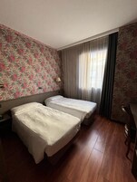 Une chambre d’hôtel avec deux lits, un papier peint à motifs floraux, un plancher en bois et une fenêtre avec des rideaux.