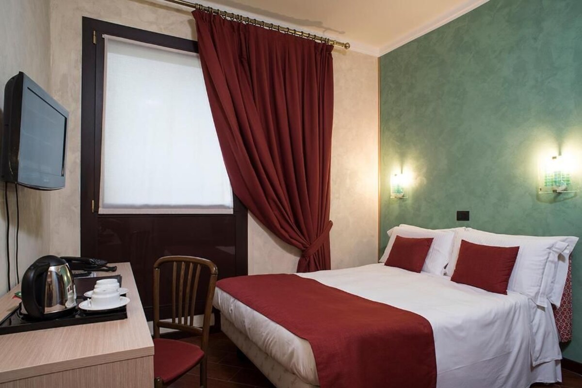 A hotel room with a bed, a desk with a kettle, a chair, a television, and a window with curtains.