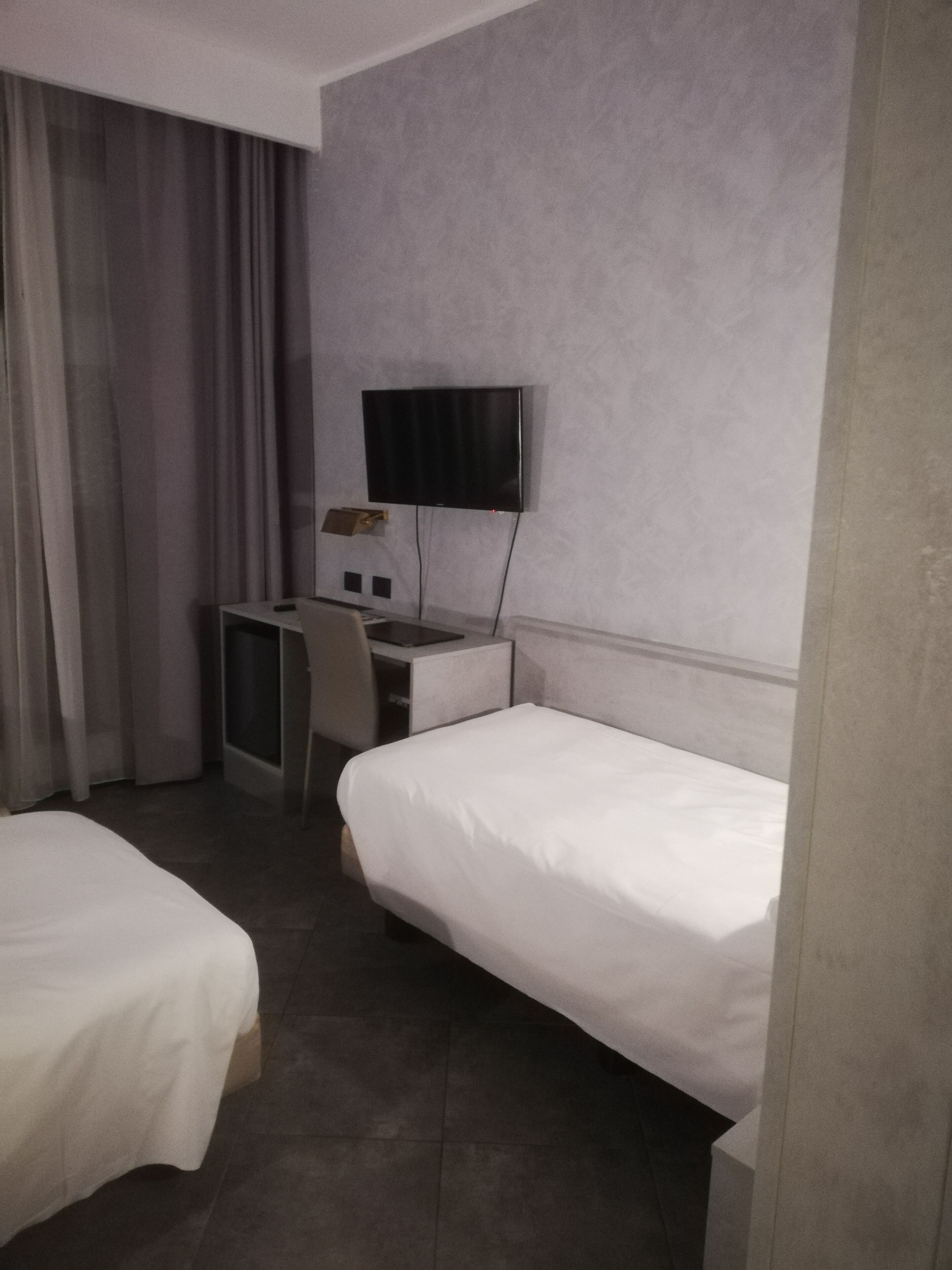 A hotel room with two single beds, a desk with a chair, a TV mounted on the wall, and a window with curtains.