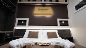 A hotel room with a bed, two bedside tables, a nightstand, a wall-mounted shelf with framed pictures, and a sign above the bed. - Hotel Plaza Torino (Turin)