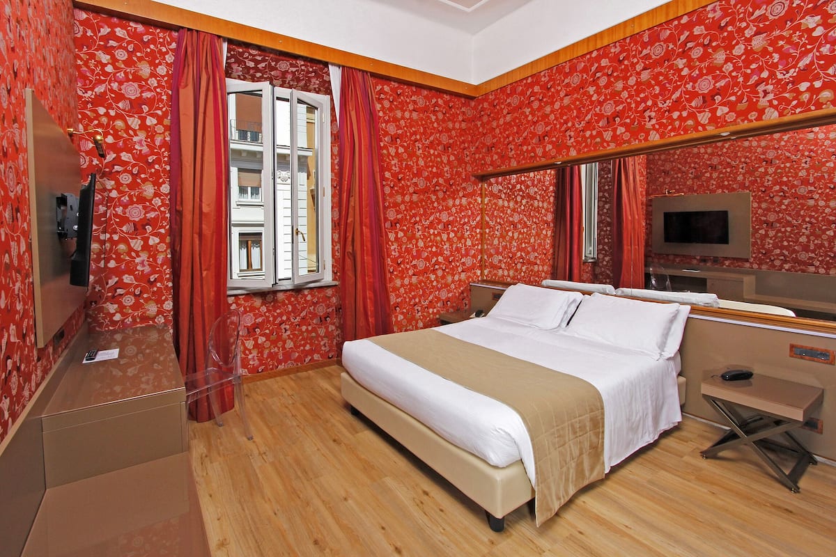 Una habitación de hotel con cama, escritorio, televisor y ventana con cortinas.