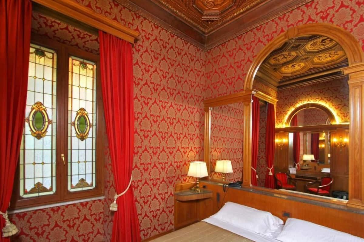 Una habitación de hotel con papel tapiz ornamentado, un espejo grande y una cama con cubrecama blanco.